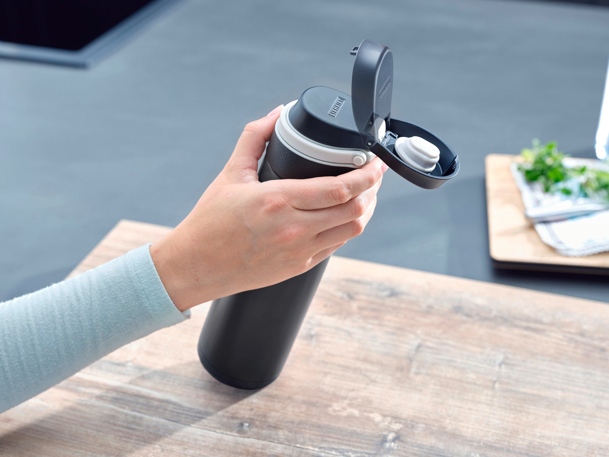 Ein schwarzer Isolierbecher Flip mit 600 ml Fassungsvermögen wird auf einem Holztisch verwendet. Eine Hand öffnet den Deckel, während im Hintergrund Küchenutensilien zu sehen sind.