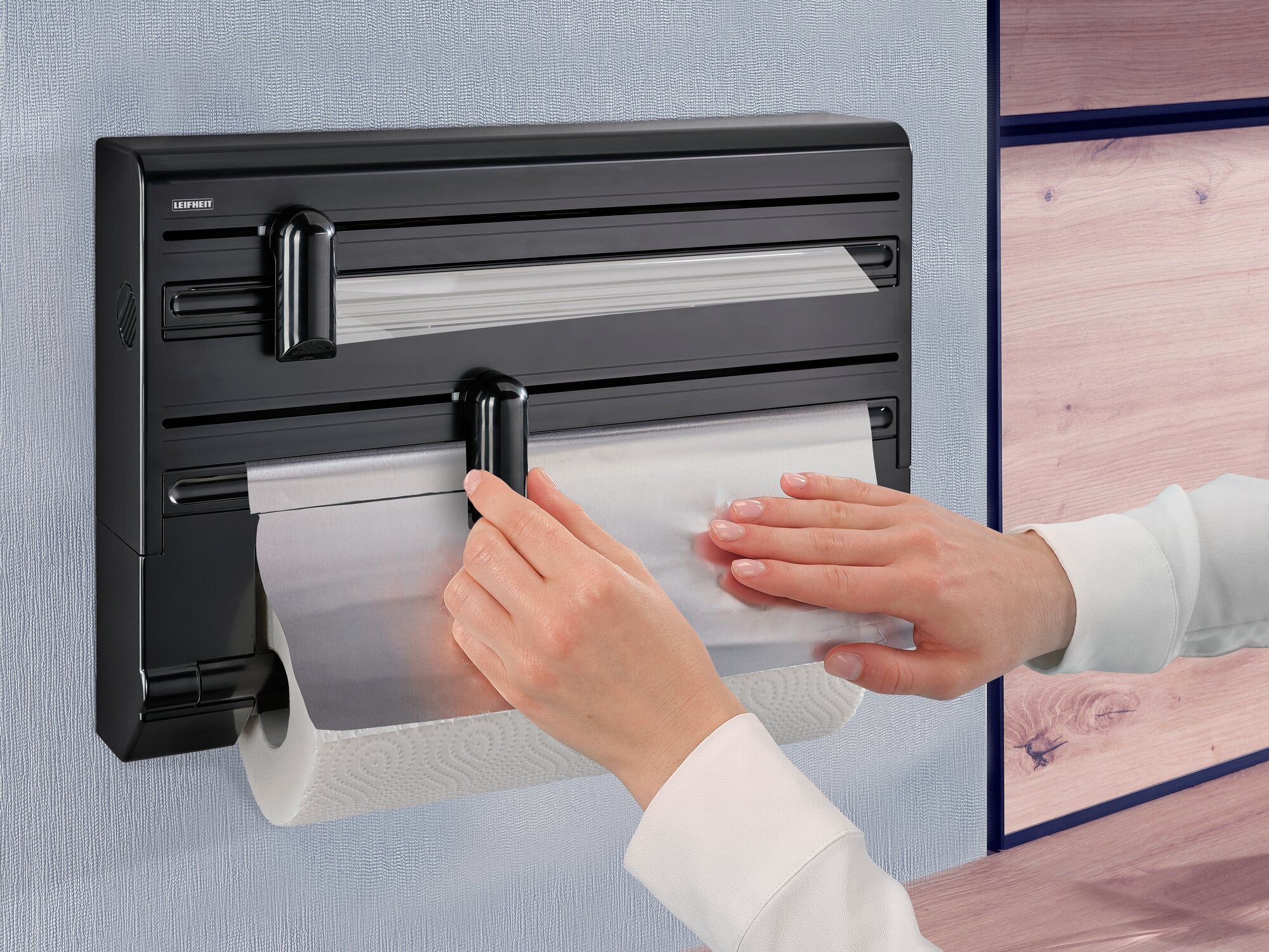 Wandrollenhalter Parat Black von Leifheit in Anwendung. Eine Person zieht Folie aus dem Halter, der an einer Wand montiert ist. Darunter befindet sich eine Küchenpapierrolle.