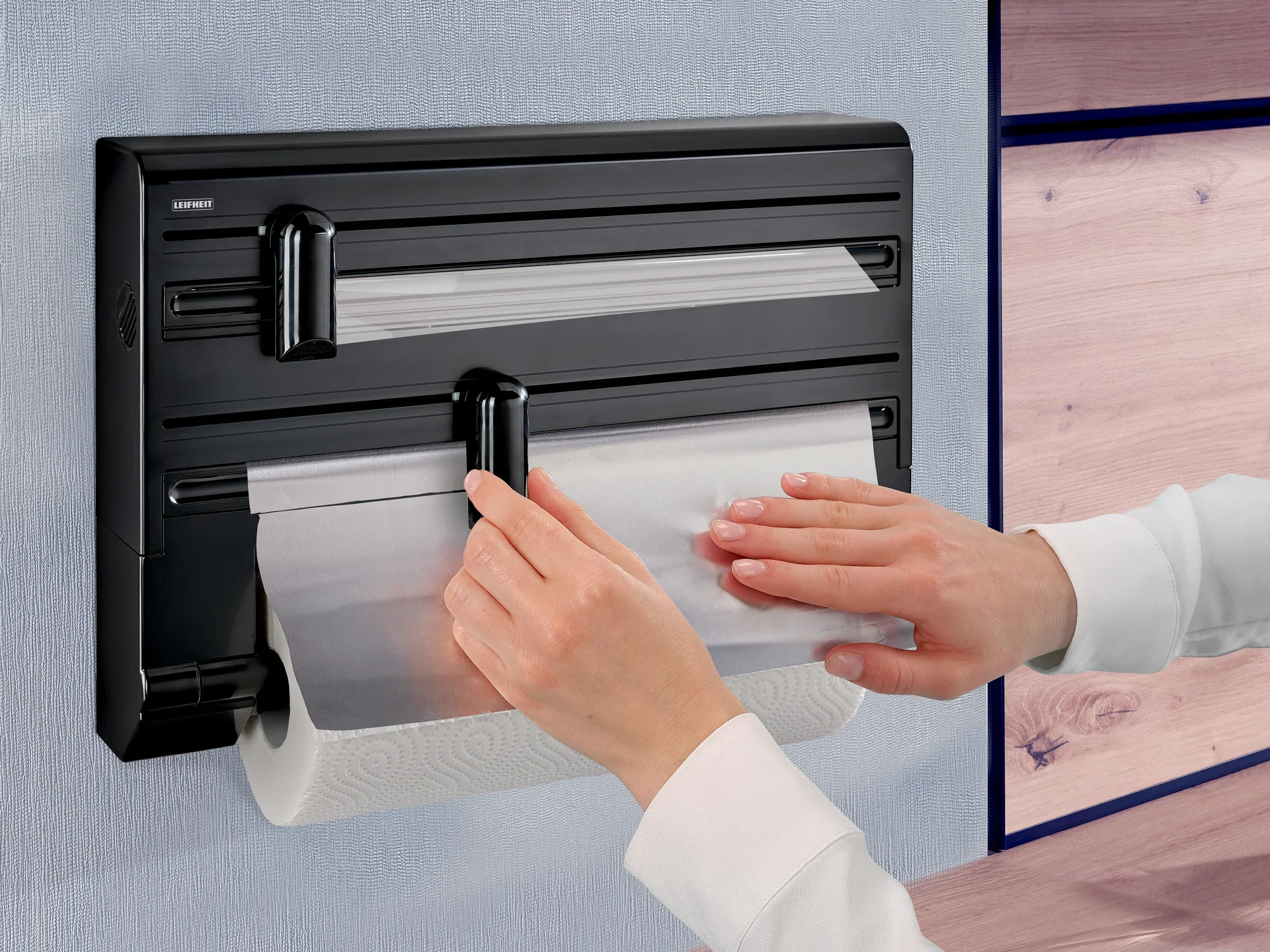 Wandrollenhalter Parat Black von Leifheit in Anwendung. Eine Person zieht Folie aus dem Halter, der an einer Wand montiert ist. Darunter befindet sich eine Küchenpapierrolle.