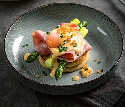 Ein Teller mit gruenem Spargel, Schinken, Eggs Benedict und Sauce Hollandaise auf Weizentoasties, garniert mit Schnittlauch und Broeseln.