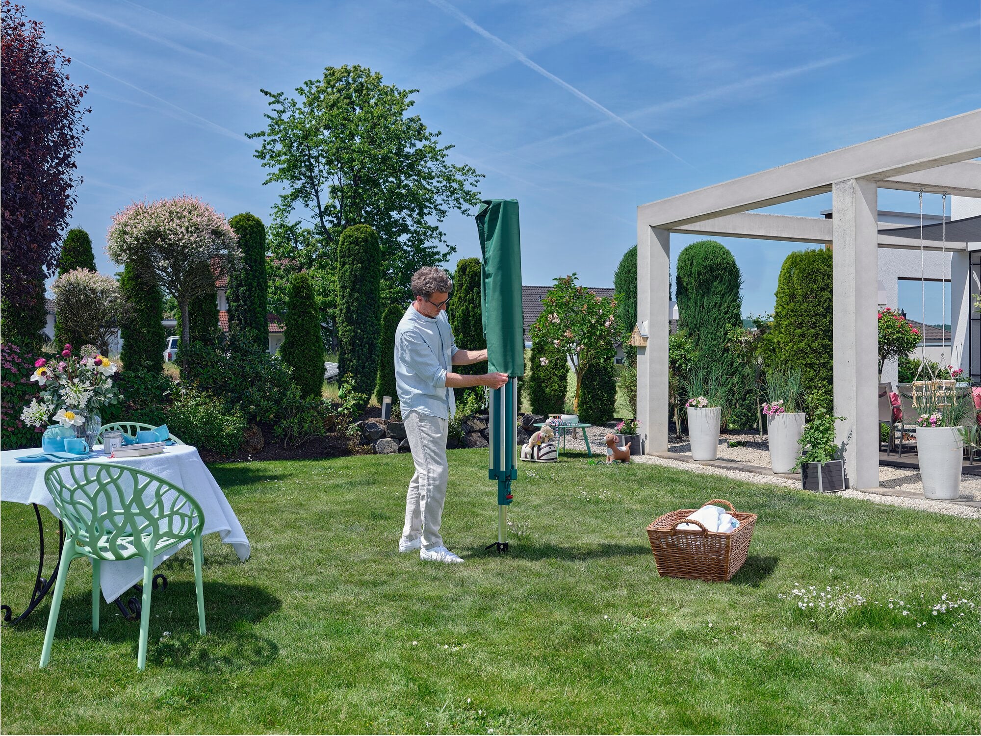 Ein Mann steht in einem gepflegten Garten und benutzt die Linomatic 500 Deluxe Wäschespinne. Umgeben von Pflanzen und einem Tisch mit Blumen, genießt er das sonnige Wetter.