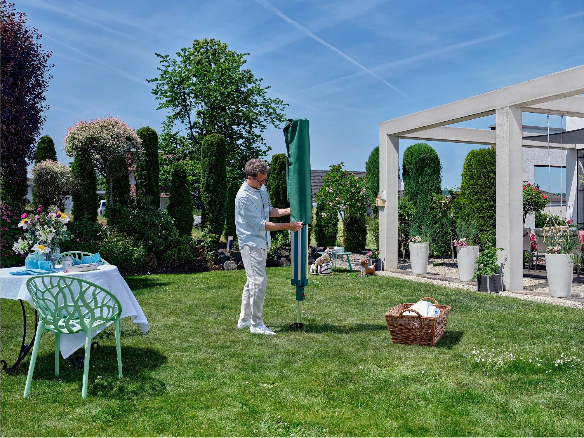 Mężczyzna stoi w zadbanym ogrodzie i korzysta z suszarki obrotowej Linomatic 500 Deluxe. Otoczony roślinami i stołem z kwiatami, cieszy się słoneczną pogodą.