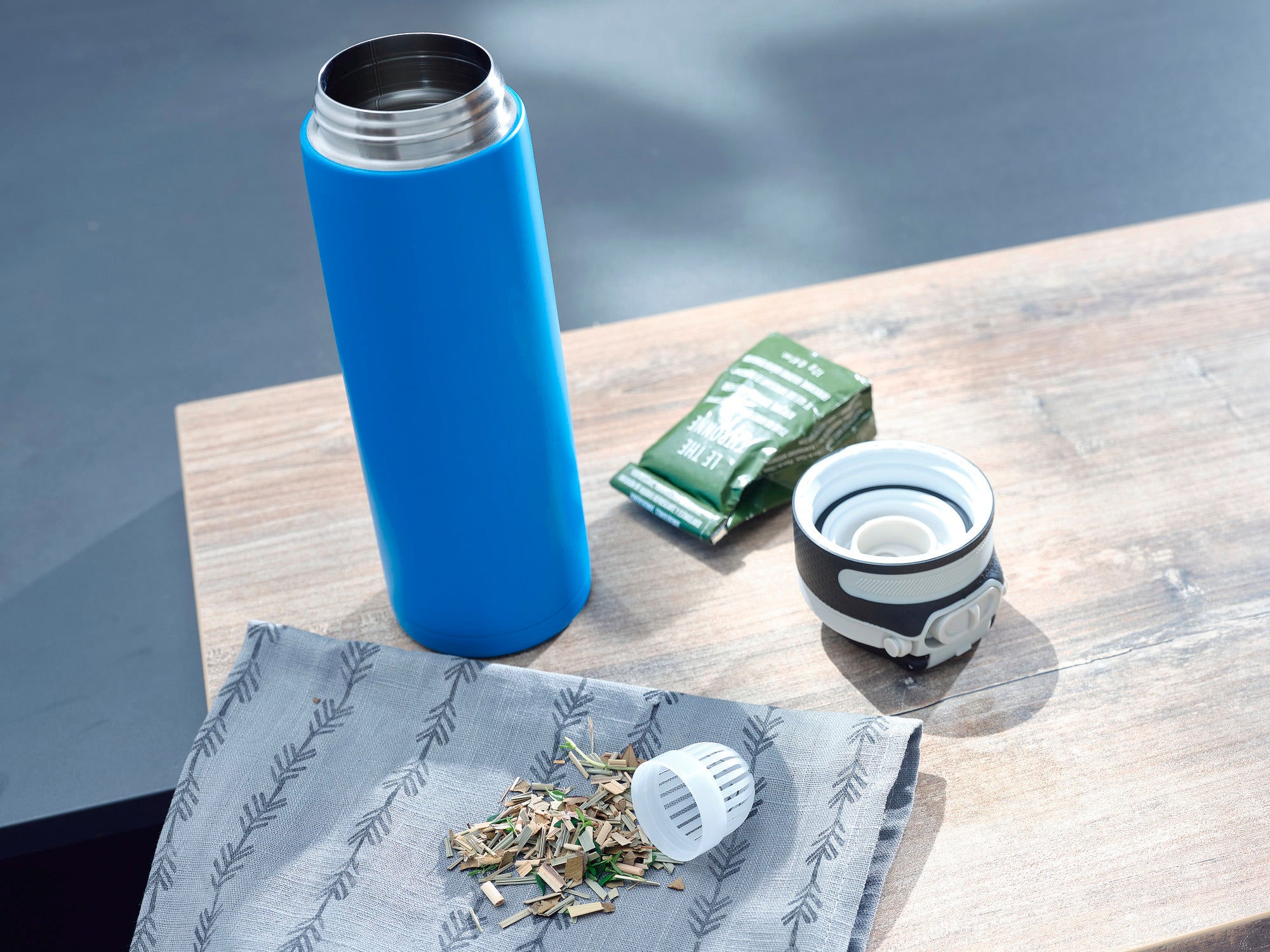 Isolierbecher Flip 600ml in Hellblau auf einem Tisch. Der Becher ist geöffnet, daneben liegen ein Teesieb, Teeblätter und eine grüne Verpackung. Ideal für unterwegs.