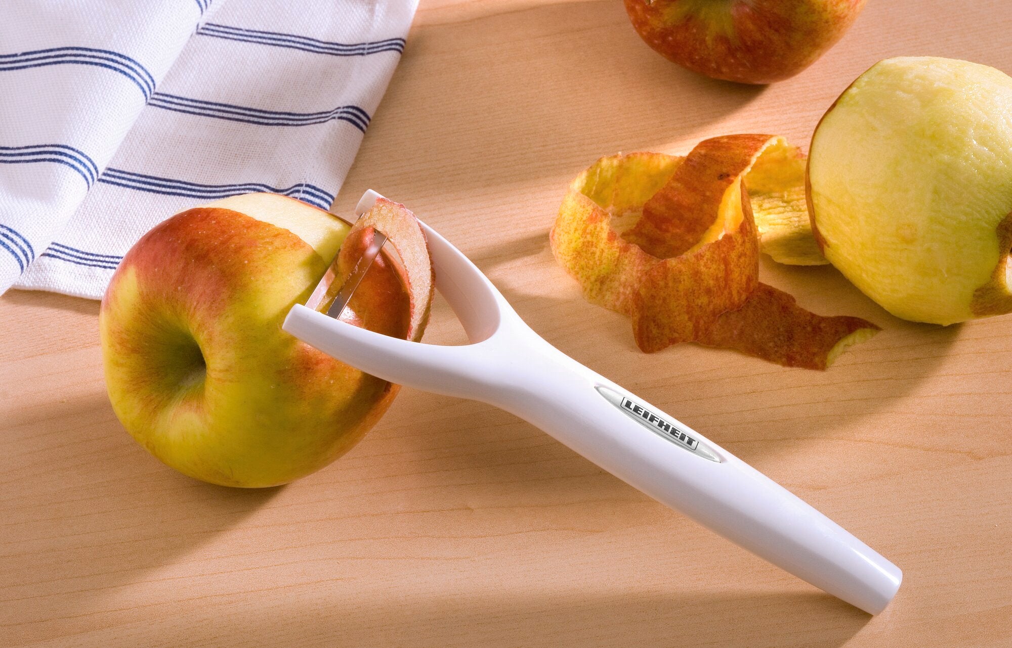Ein Leifheit Sparschäler mit weißem Griff liegt auf einem Holztisch. Er wird verwendet, um die Schale eines Apfels zu entfernen. Im Hintergrund sind weitere geschälte Äpfel zu sehen.