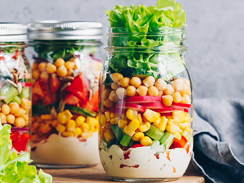 Drei Einmachglaeser mit Schichtsalat aus Kichererbsen, Mais, Paprika, Gurken und Salatblaettern, auf cremigem Dressing angerichtet.