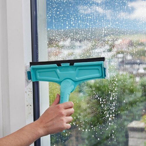 Eine Hand hält einen türkisfarbenen Fensterabzieher, der ein großes Fenster reinigt. Im Hintergrund ist eine verschwommene Stadtlandschaft zu sehen. Der Abzieher entfernt effektiv Wasser und Schmutz.
