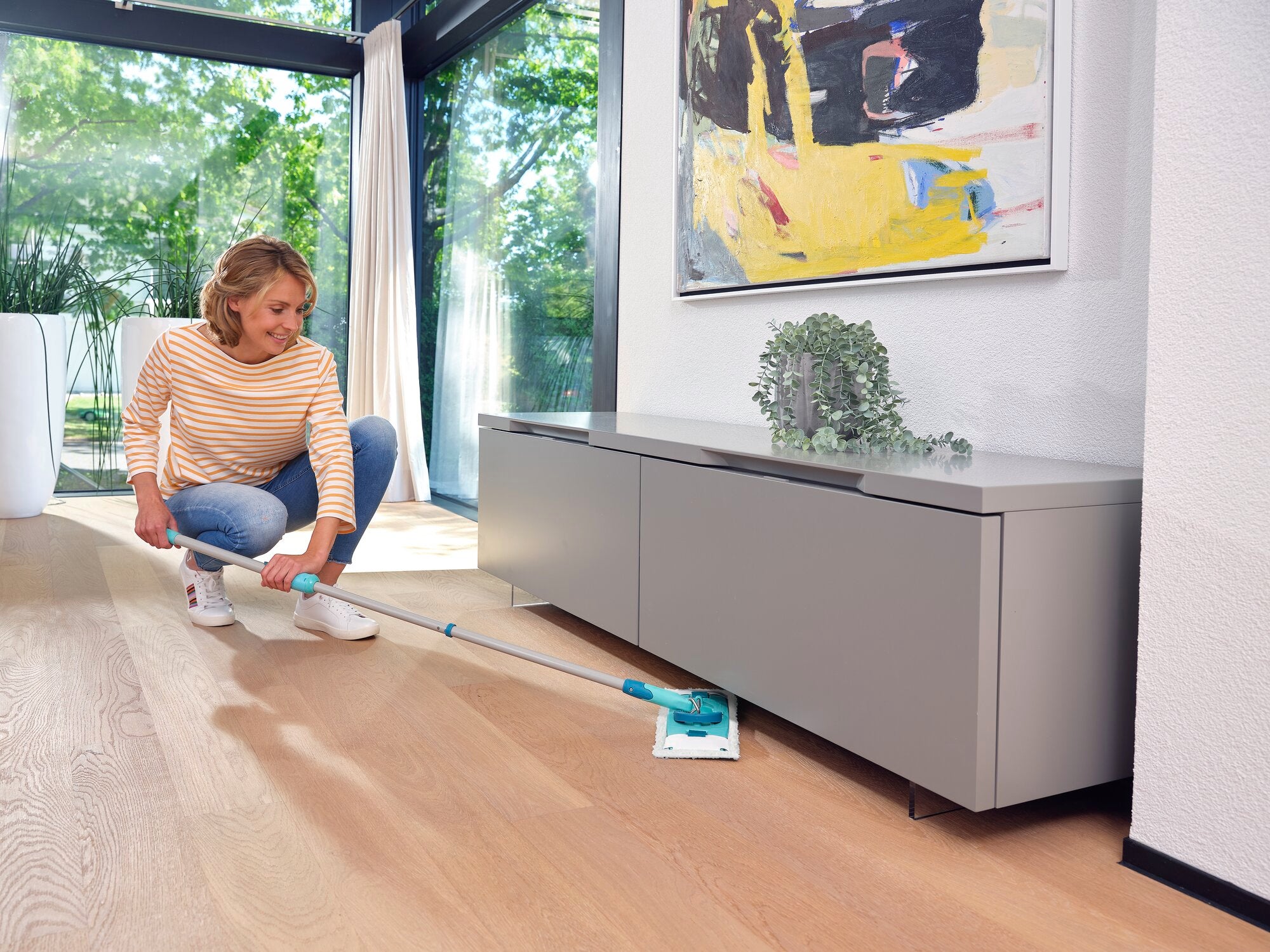 Kobieta czyści drewnianą podłogę w nowoczesnym salonie za pomocą CLEAN TWIST M Ergo. Urządzenie umożliwia łatwe czyszczenie pod meblami. Idealny do dokładnego czyszczenia.