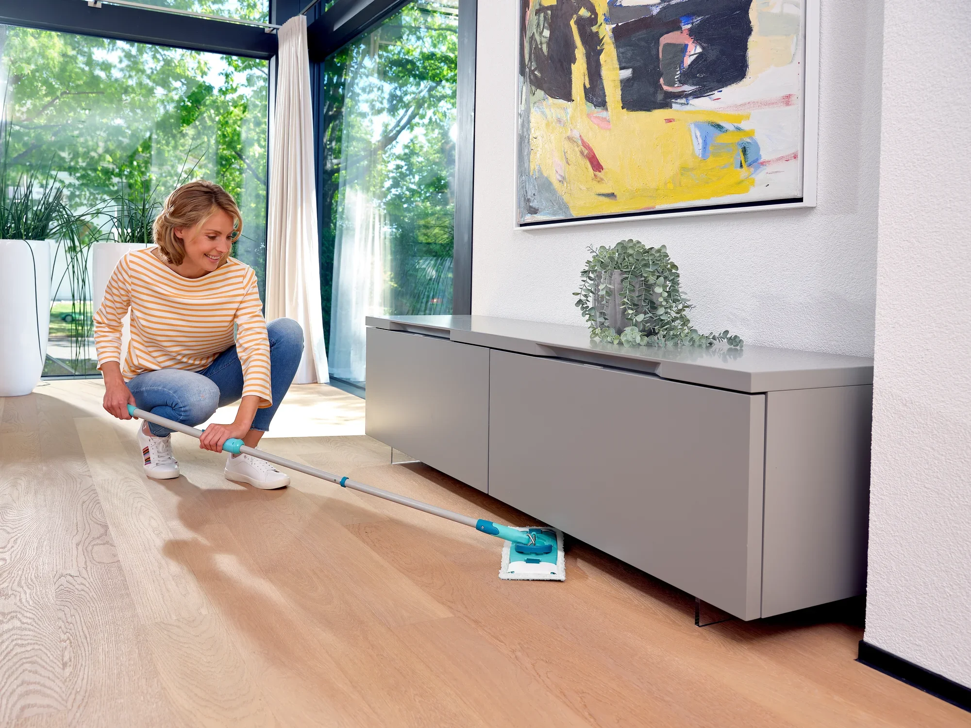 Kobieta czyści drewnianą podłogę w nowoczesnym salonie za pomocą CLEAN TWIST M Ergo. Urządzenie umożliwia łatwe czyszczenie pod meblami. Idealny do dokładnego czyszczenia.