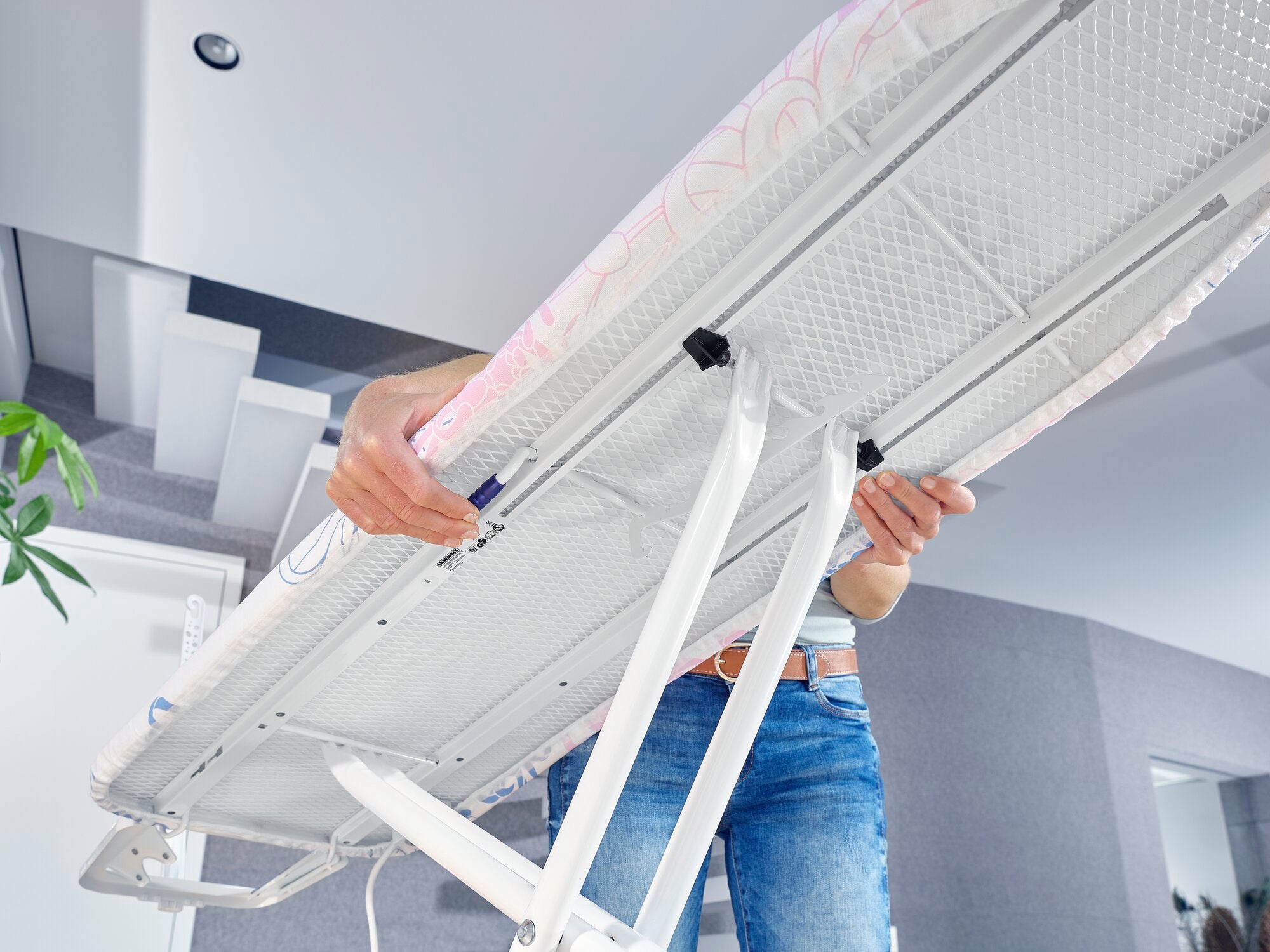 Tabla de planchar Classic M Compact Plus: una persona ajusta la altura de una tabla de planchar. La tabla tiene un bastidor blanco estable y una superficie de planchado estampada.