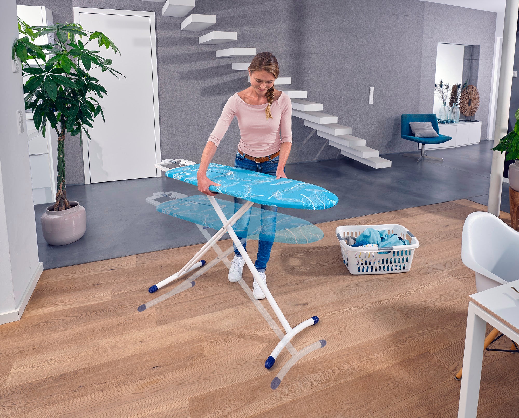 Eine Frau bügelt auf einem Leifheit Air Board M Solid Bügeltisch in einem modernen Wohnzimmer. Der Tisch ist leicht und hat eine blaue Bügelfläche. Im Hintergrund steht ein Wäschekorb.