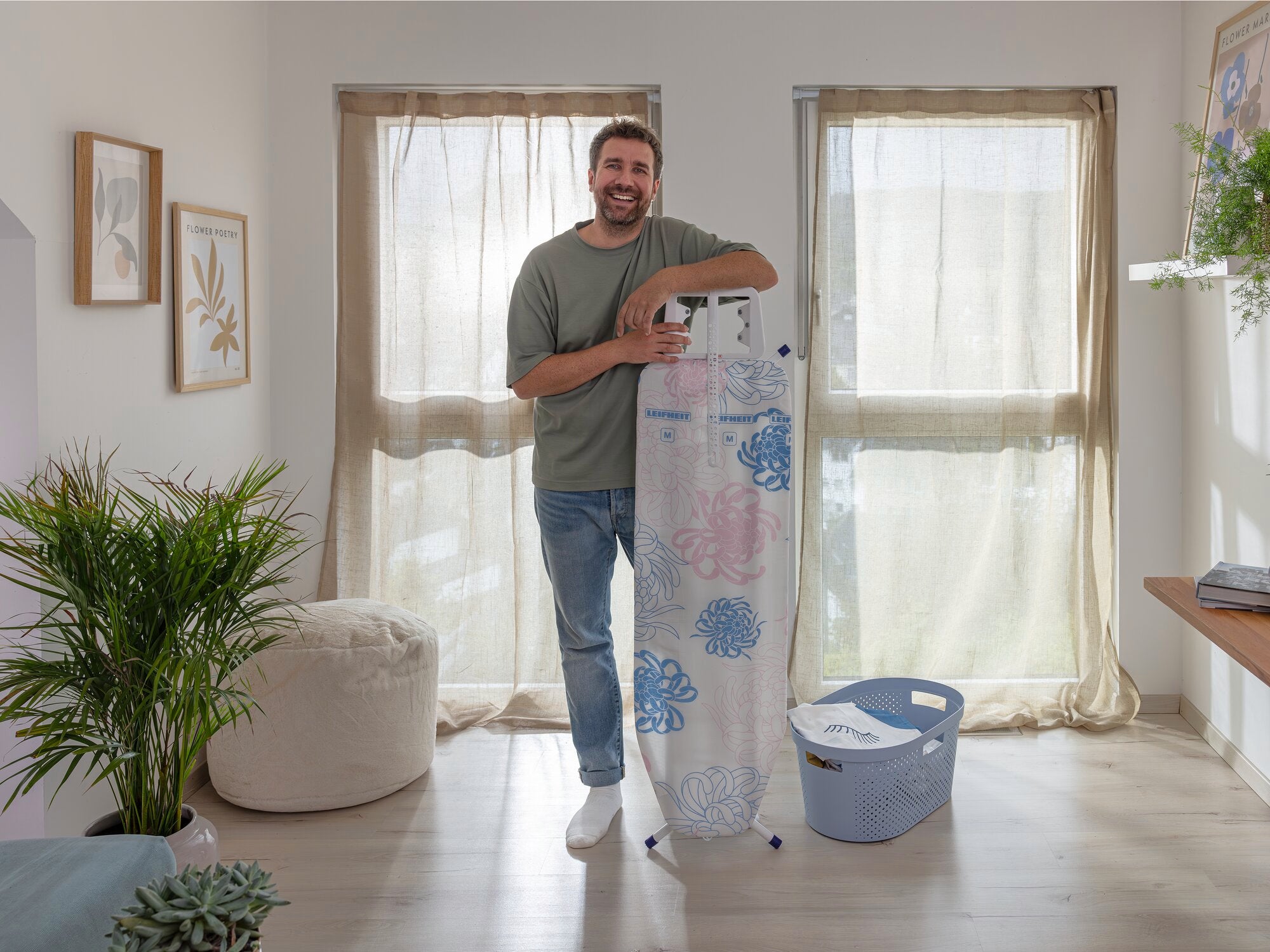 Un homme sourit à côté d'une table à repasser Classic M Basic Plus dans une pièce lumineuse. La table est recouverte d'une housse colorée. À l'arrière-plan, on aperçoit deux grandes fenêtres et un panier à linge.