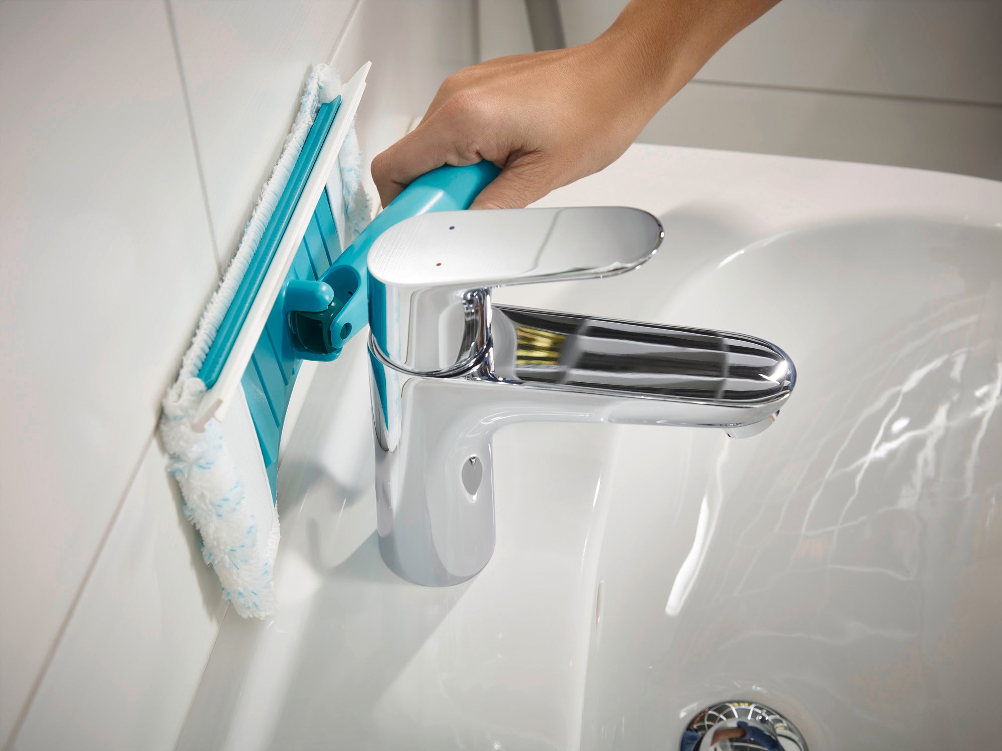 A Flexipad tile and tub squeegee with a telescopic handle is being used on a bathroom wall. The squeegee has a blue pad and is guided by one hand to clean the tiles.