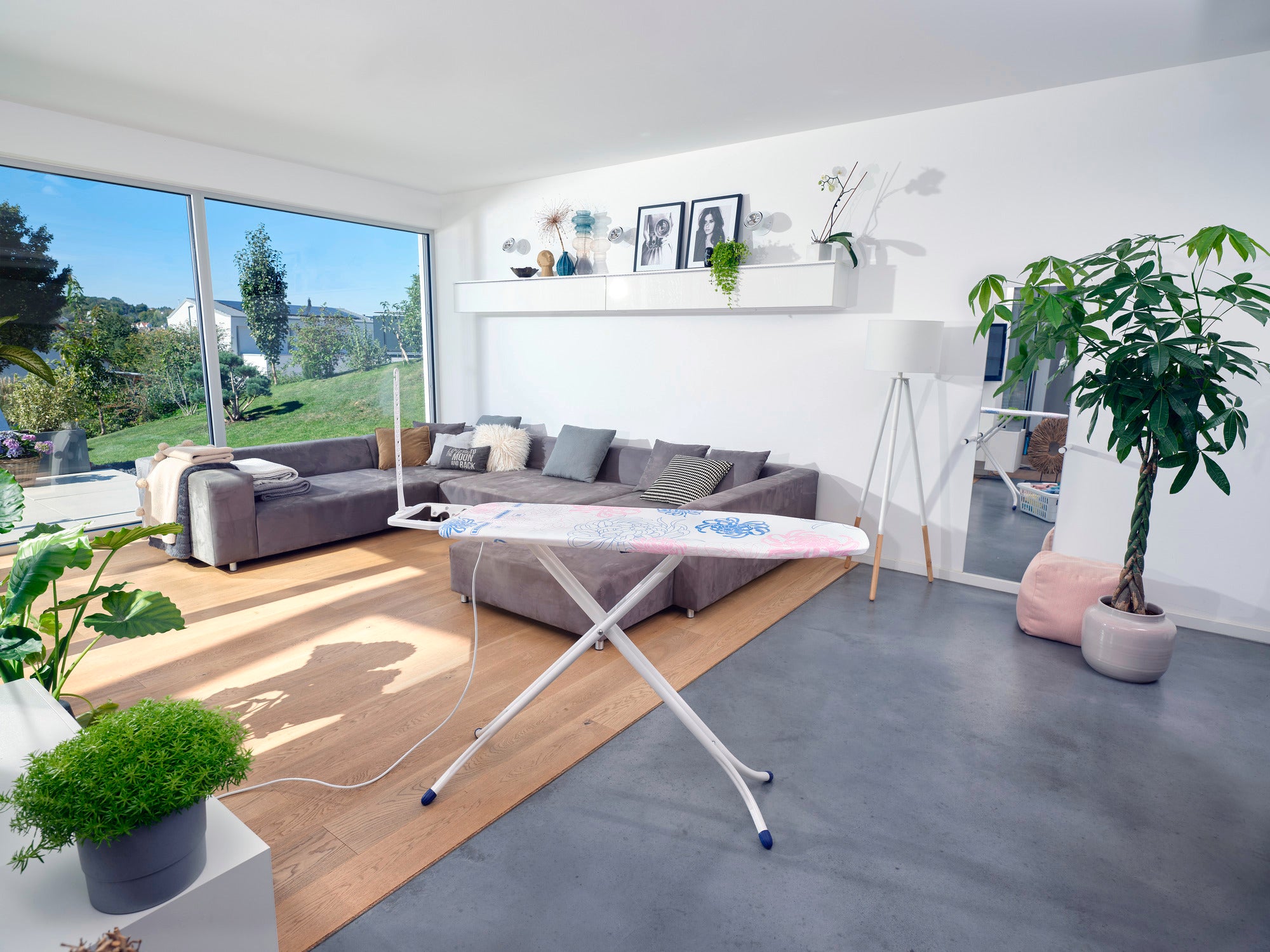 Bügeltisch Classic M Compact Plus in einem modernen Wohnzimmer mit großen Fenstern, die viel Tageslicht hereinlassen. Der Tisch steht auf einem grauen Boden, umgeben von Pflanzen und einem Sofa.