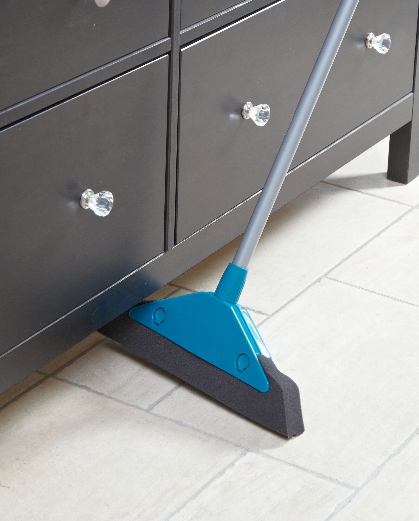 Balai en mousse en action : un balai en mousse bleu nettoie efficacement le sol sous une armoire noire avec des poignées brillantes sur du carrelage clair.