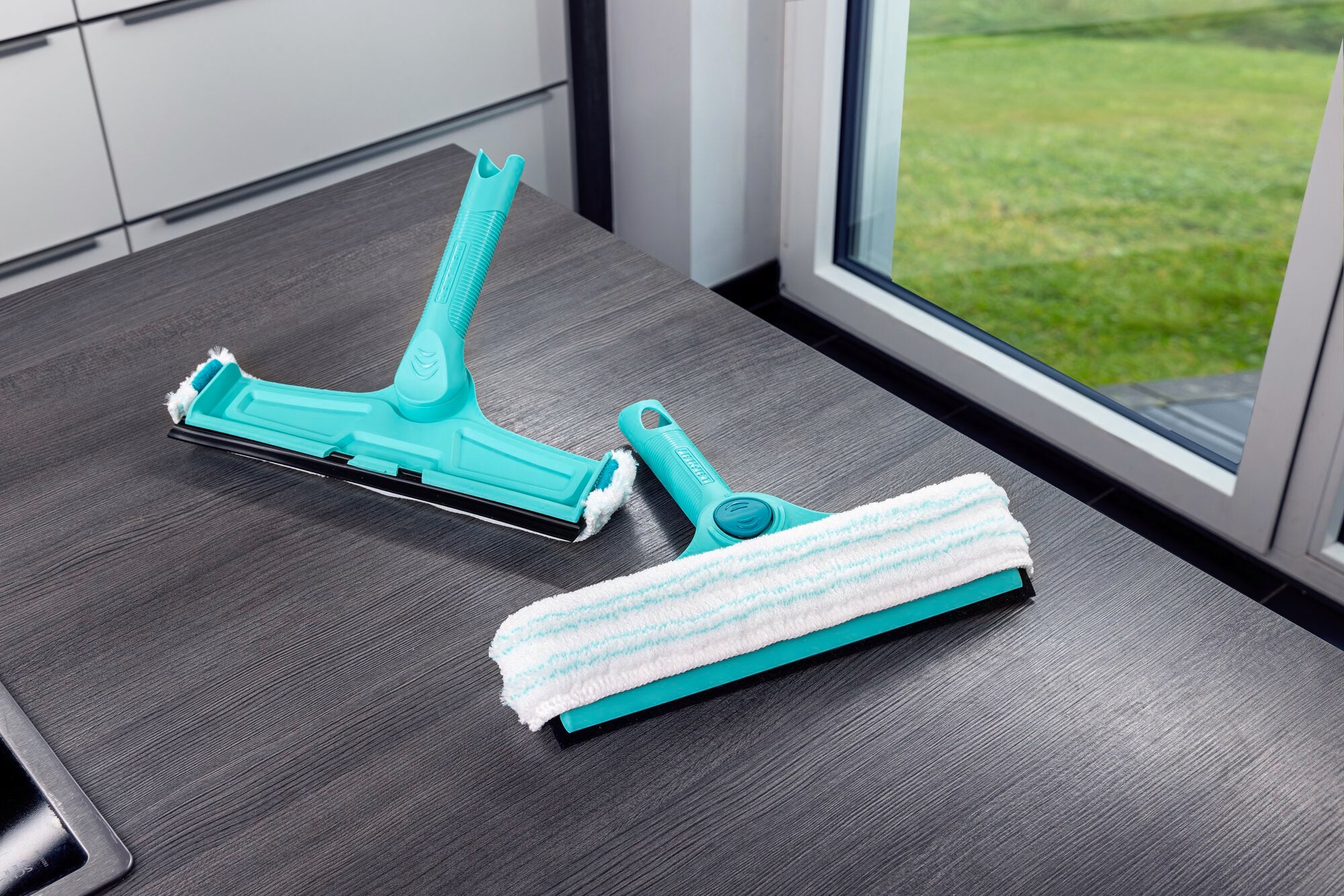 Zwei türkisfarbene Fenster- und Rahmenreiniger 4in1 liegen auf einem Tisch vor einem Fenster. Der eine hat einen Mikrofaserbezug, der andere eine glatte Kante. Ideal für saubere Fenster.