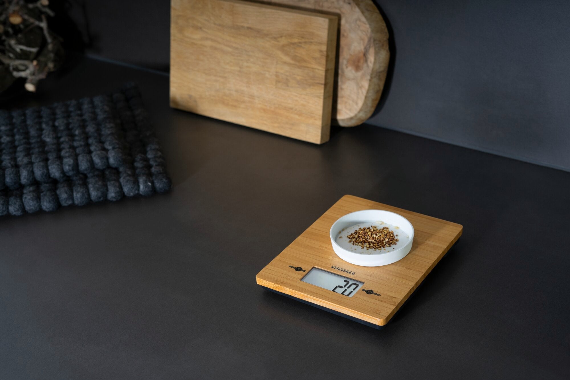 Digitale keukenweegschaal van bamboe op een donker werkblad. Een kleine kom met zaden staat erop. Op de achtergrond zijn houten snijplanken te zien. Ideaal voor nauwkeurig wegen in de keuken.