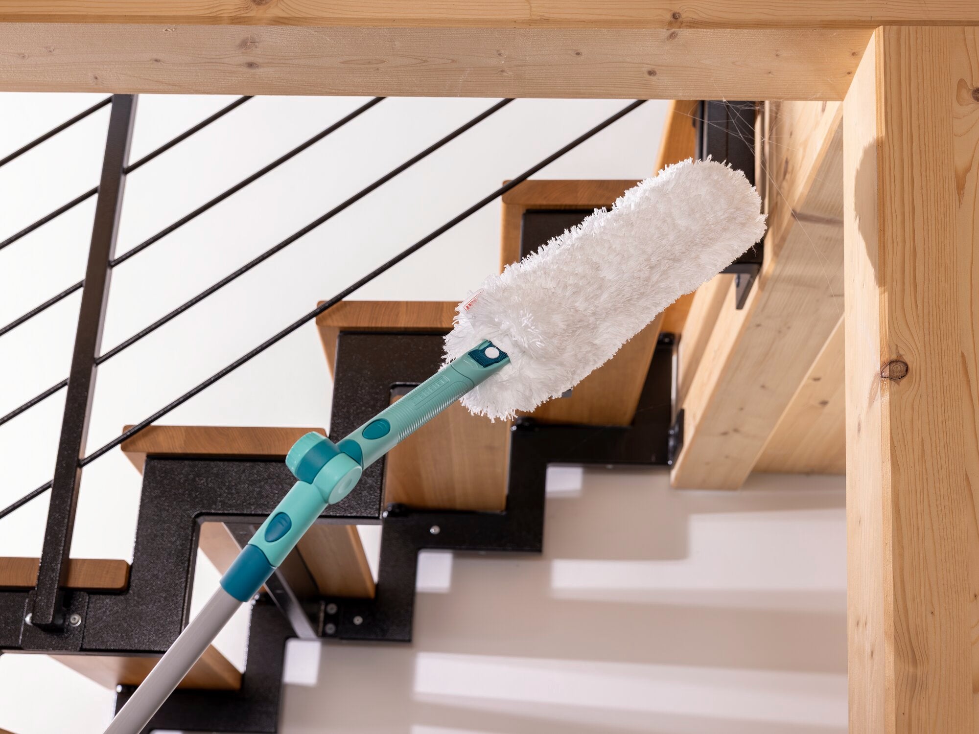 Een SUPERDUSTER XL plumeau wordt gebruikt om moeilijk bereikbare plaatsen op een houten trap schoon te maken. De plumeau heeft een lange steel en een pluizig oppervlak.