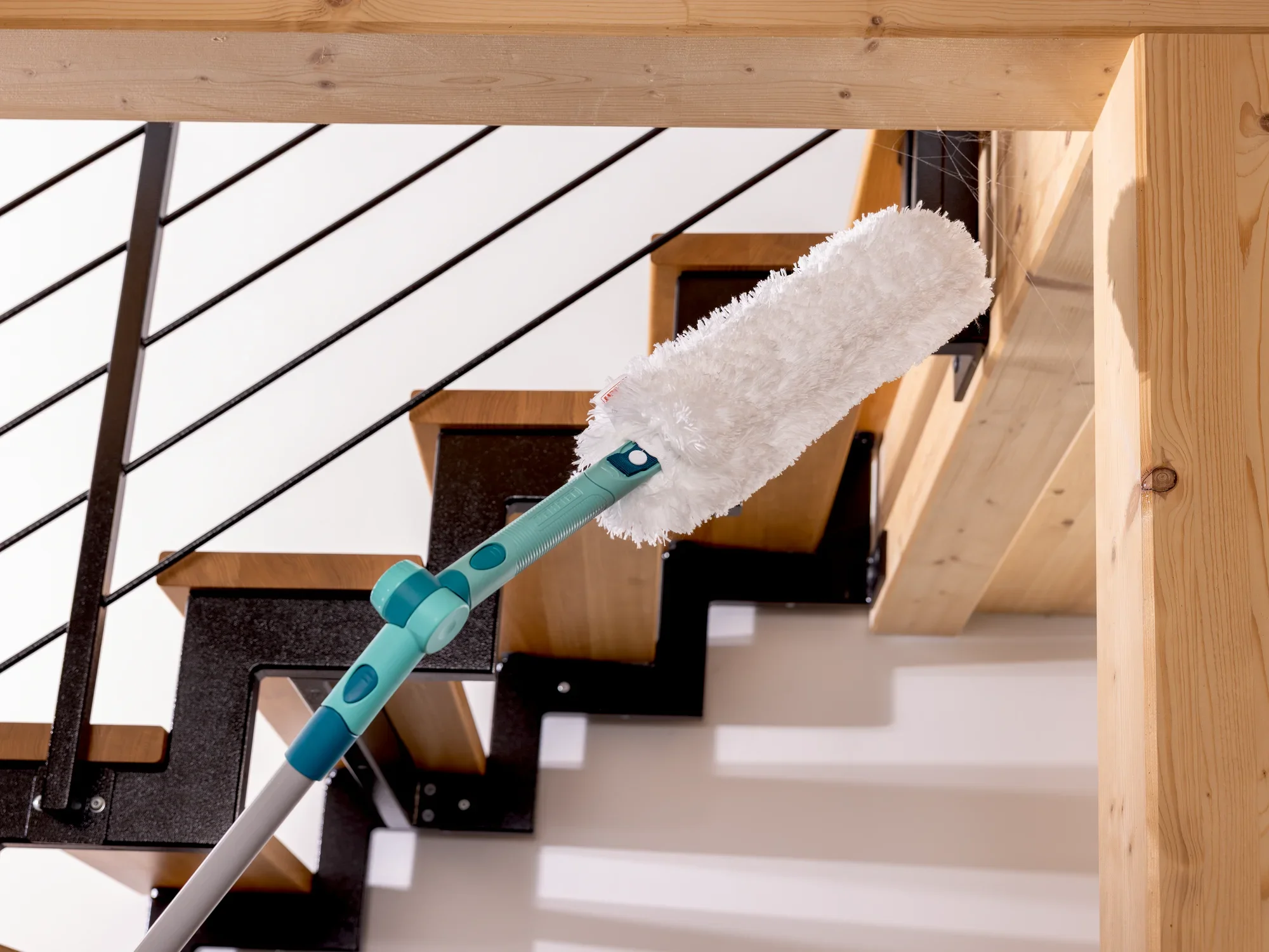 Een SUPERDUSTER XL plumeau wordt gebruikt om moeilijk bereikbare plaatsen op een houten trap schoon te maken. De plumeau heeft een lange steel en een pluizig oppervlak.