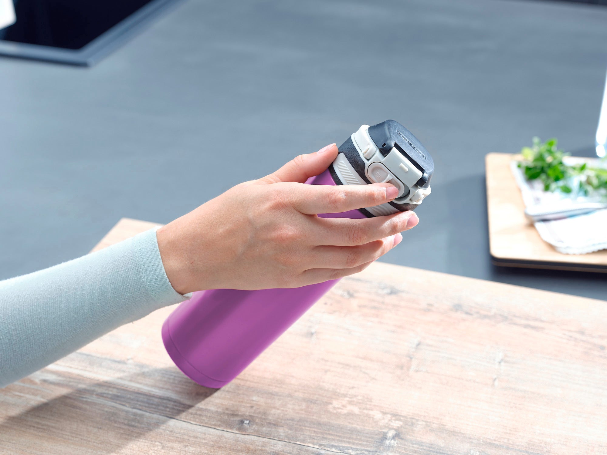 Flip 600 ml insulated mug in purple on a table. A hand opens the lid of the mug. Kitchen utensils and herbs can be seen in the background.