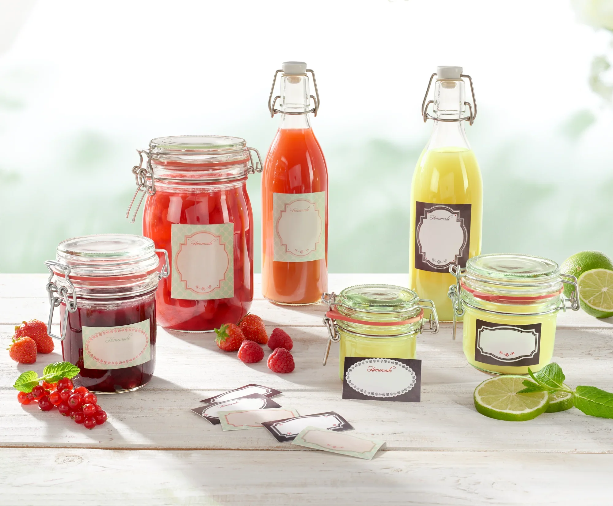 Różne szklane pojemniki z czerwonymi i żółtymi napojami i dżemami, ozdobione mieszanymi dekoracyjnymi etykietami. Świeże jagody i limonki leżą obok nich na białym stole.