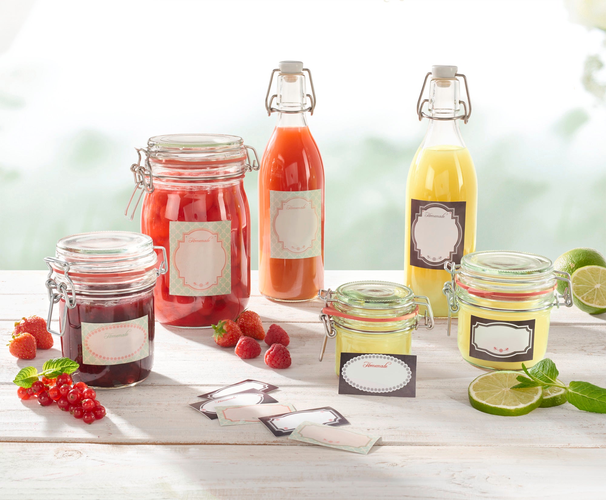 Verschiedene Glasbehälter mit roten und gelben Getränken sowie Marmeladen, dekoriert mit gemischten Deko-Etiketten. Frische Beeren und Limetten liegen daneben auf einem weißen Tisch.