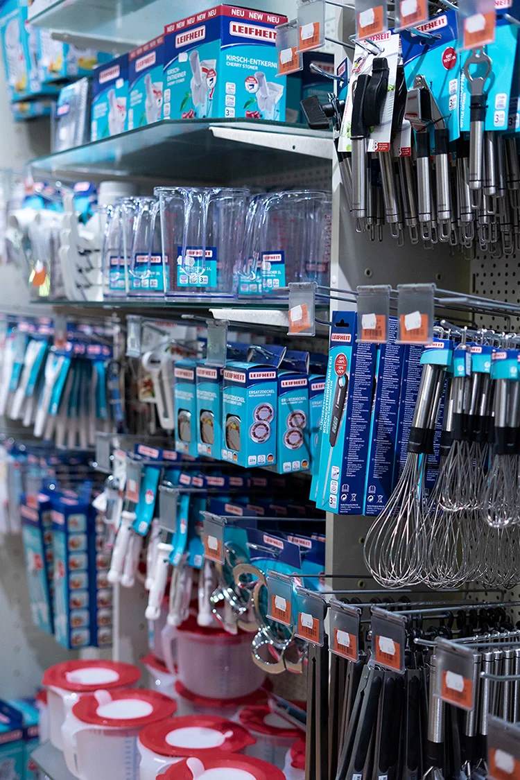 Regale in einem Geschaeft mit Kuechenutensilien wie Scheren, Schneebesen, Messbechern und Produkten in blauen Verpackungen.