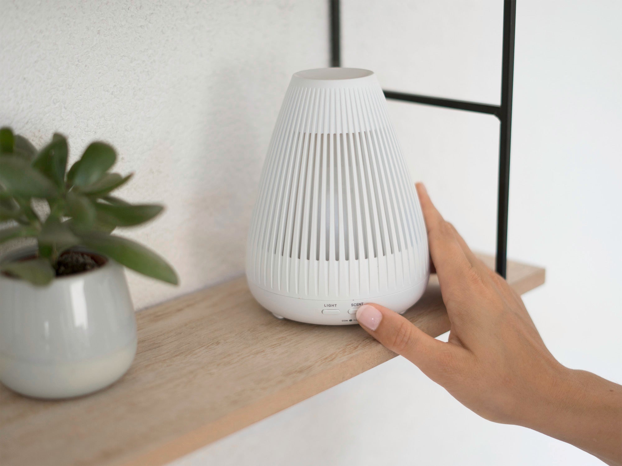 Ein weißer Design Aroma Diffuser Bari steht auf einem Holzregal neben einer kleinen Pflanze. Eine Hand berührt sanft das Gerät, um es zu aktivieren. Ideal für ein stilvolles Ambiente.