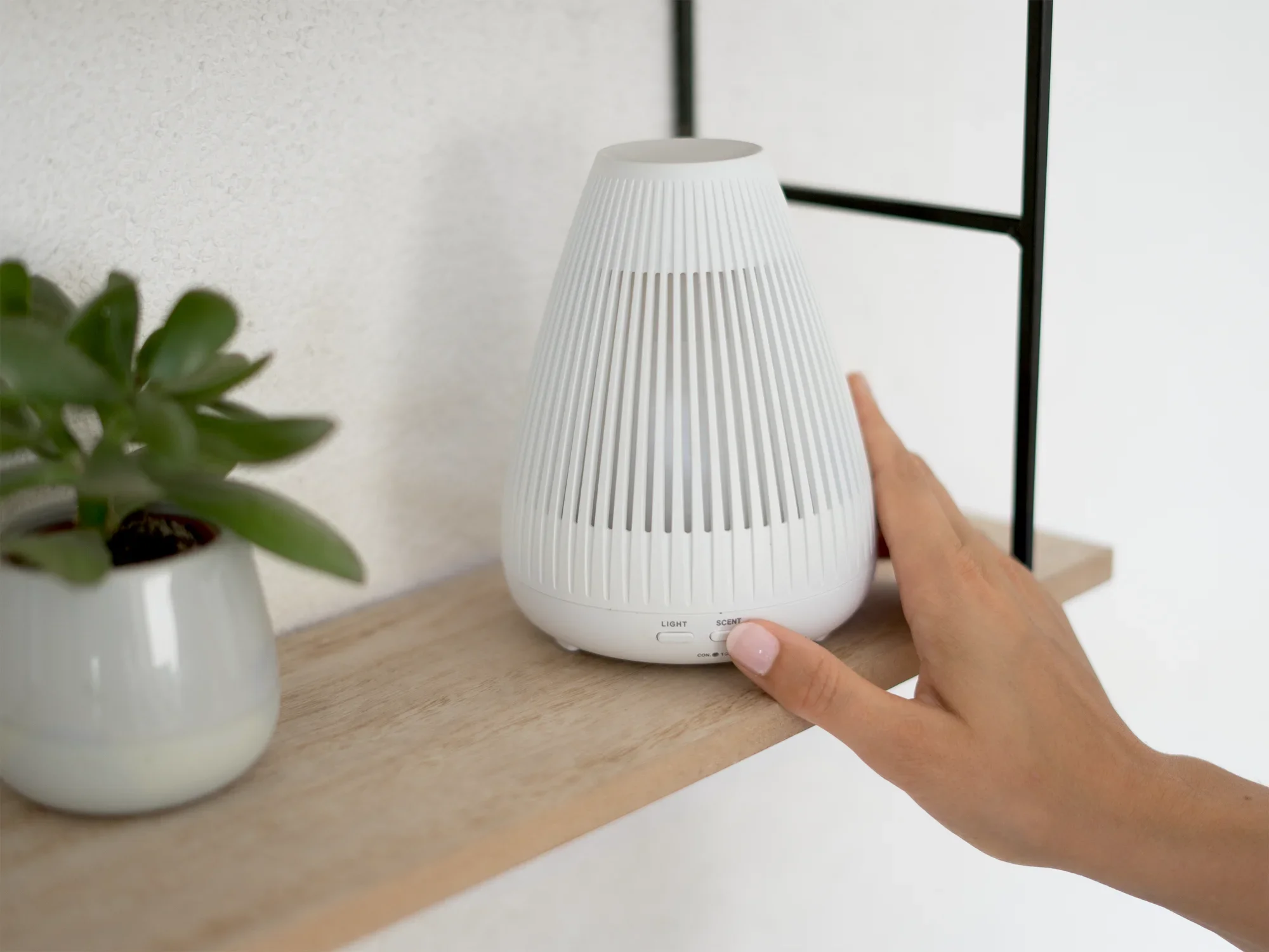 Un diffuseur d'arômes design Bari blanc est posé sur une étagère en bois à côté d'une petite plante. Une main touche délicatement l'appareil pour l'activer. Idéal pour créer une ambiance élégante.