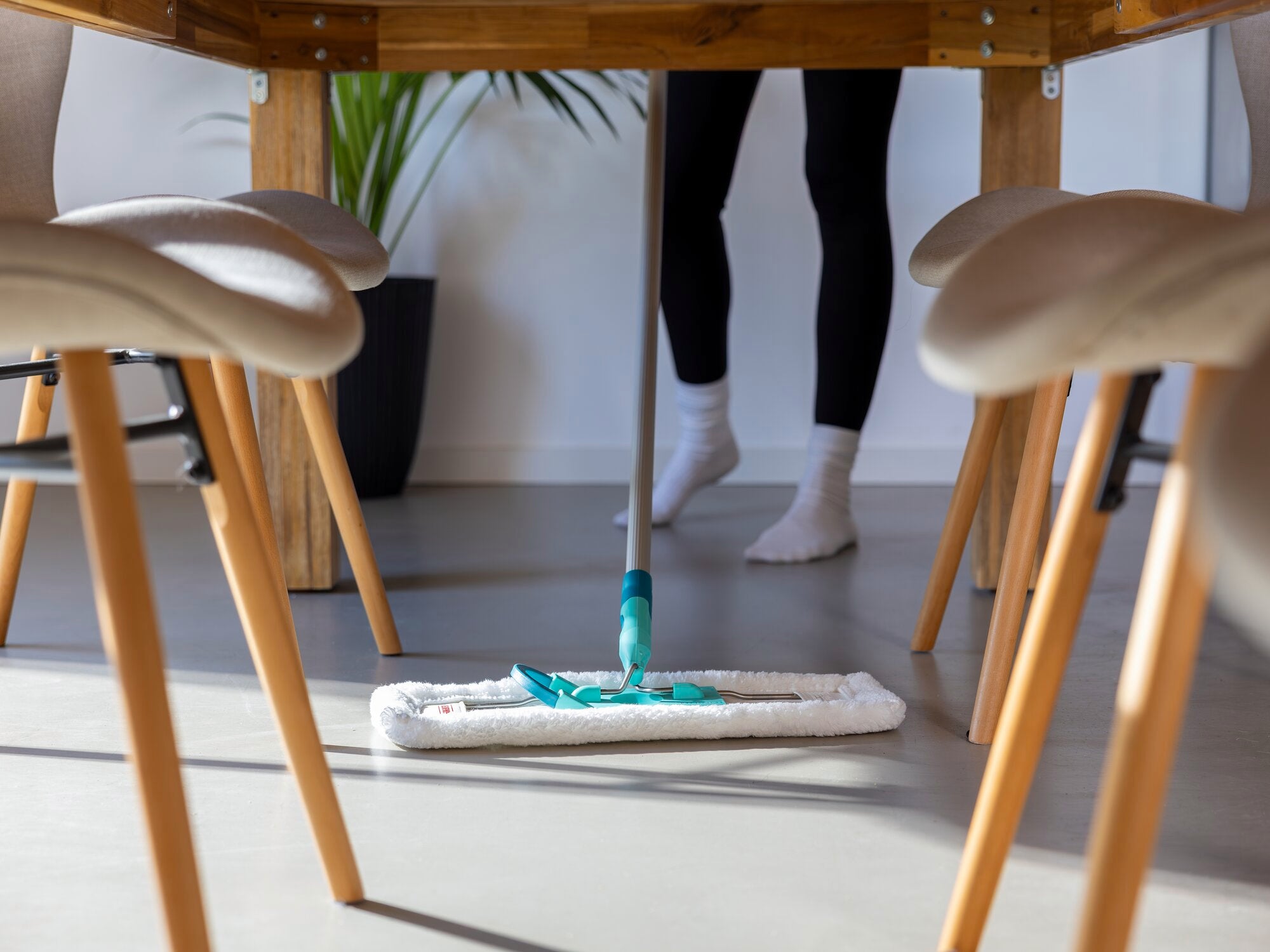 Ein Staubwischer SUPERDUSTER Floor XL wird unter einem Tisch verwendet. Eine Person wischt den Boden mit dem Staubwischer, während sie weiße Socken trägt. Im Hintergrund ist eine Pflanze sichtbar.