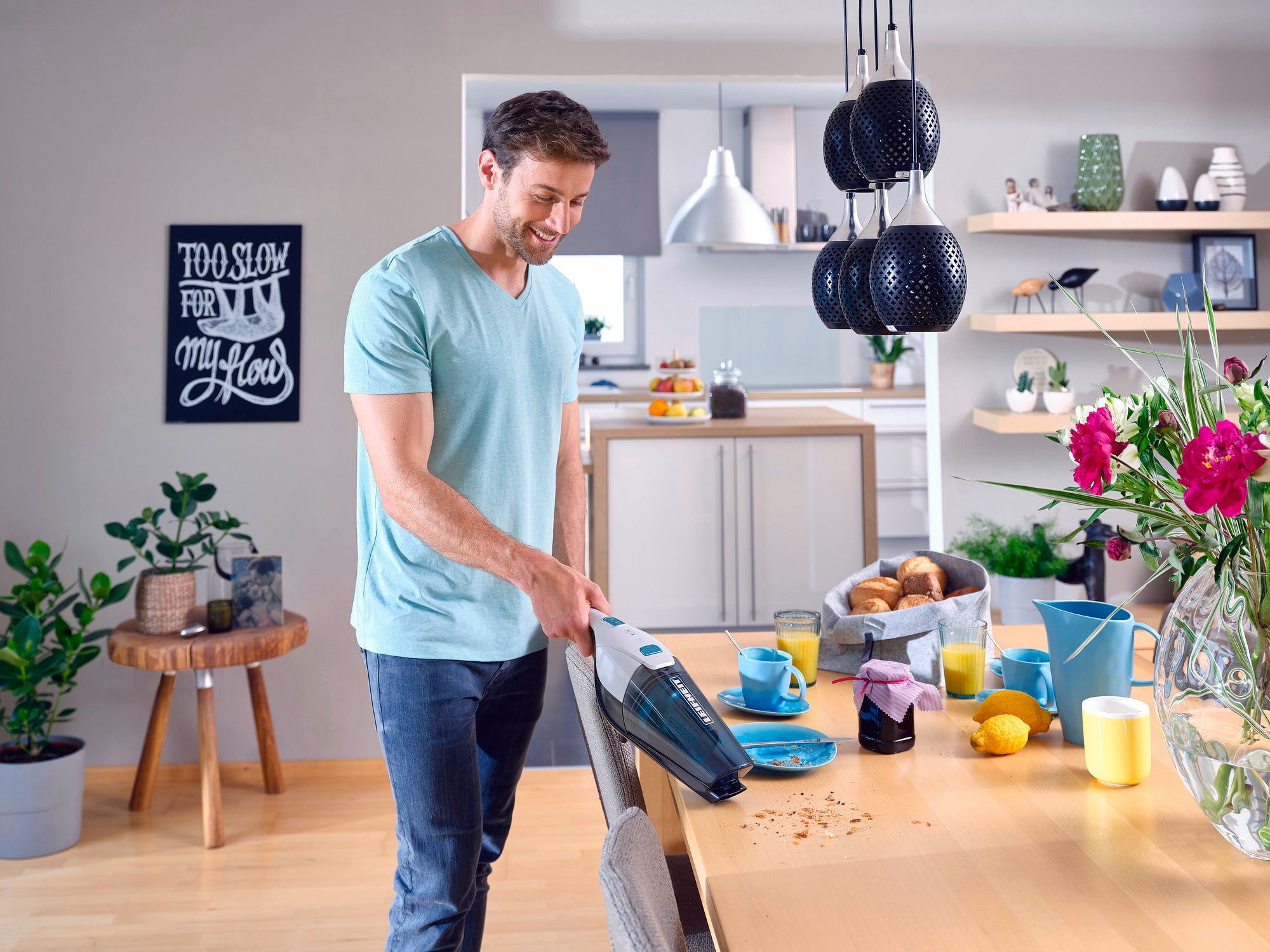 Ein Mann benutzt den Akku-Staubsauger Rotaro PowerVac, um Krümel von einem Tisch in einer modernen Küche zu entfernen. Im Hintergrund sind Pflanzen und Küchenutensilien zu sehen.