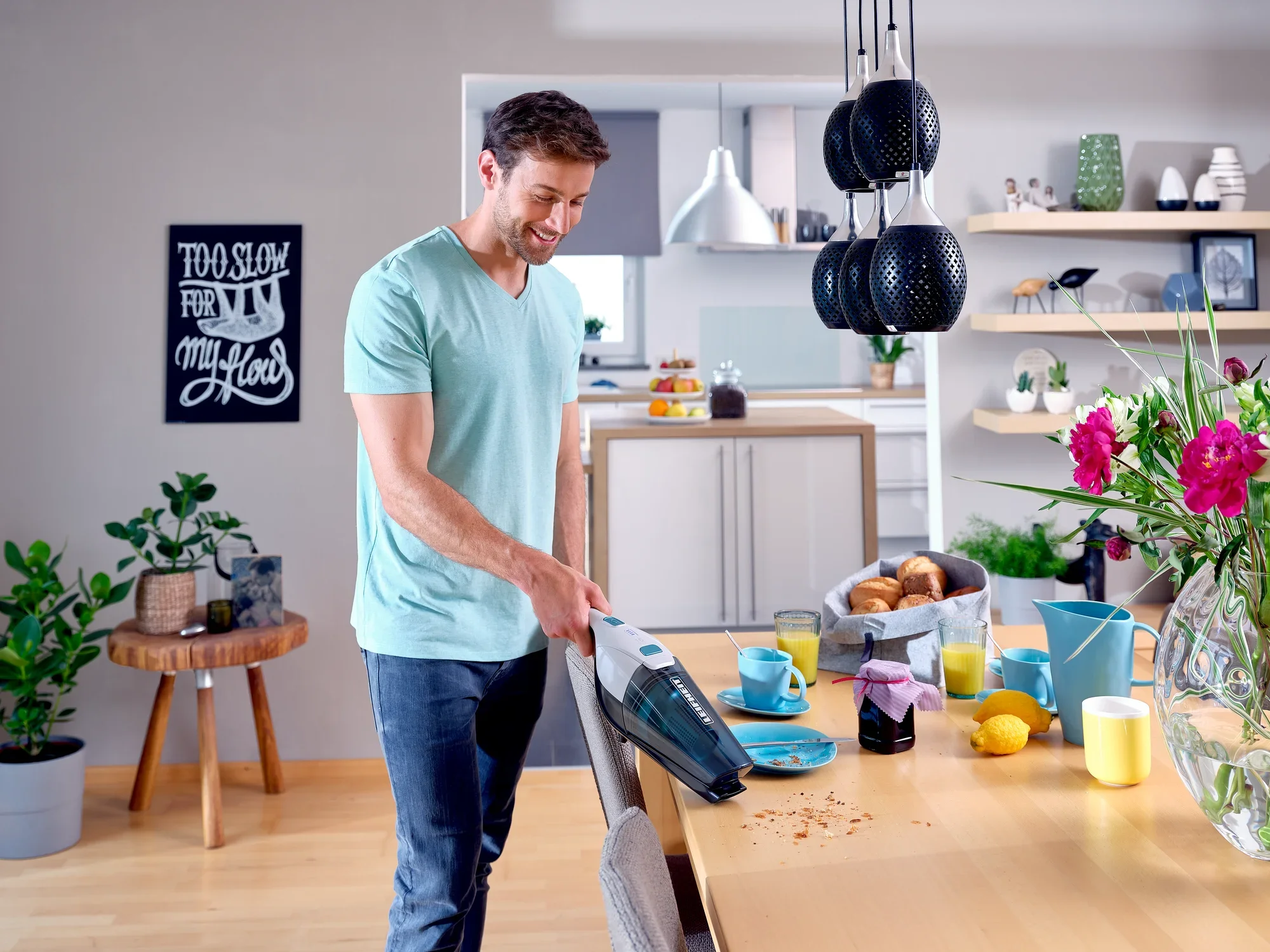 Un homme utilise l'aspirateur sans fil Rotaro PowerVac pour enlever des miettes sur une table dans une cuisine moderne. On aperçoit des plantes et des ustensiles de cuisine à l'arrière-plan.