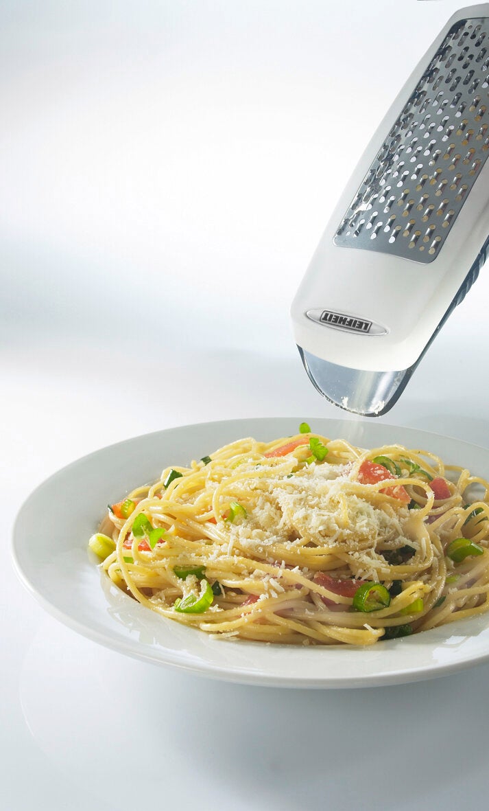 Eine Parmesanreibe wird über einem Teller Spaghetti verwendet, um frischen Parmesan zu reiben. Die Reibe ist aus Edelstahl und zeigt ihre Effizienz in der Anwendung.