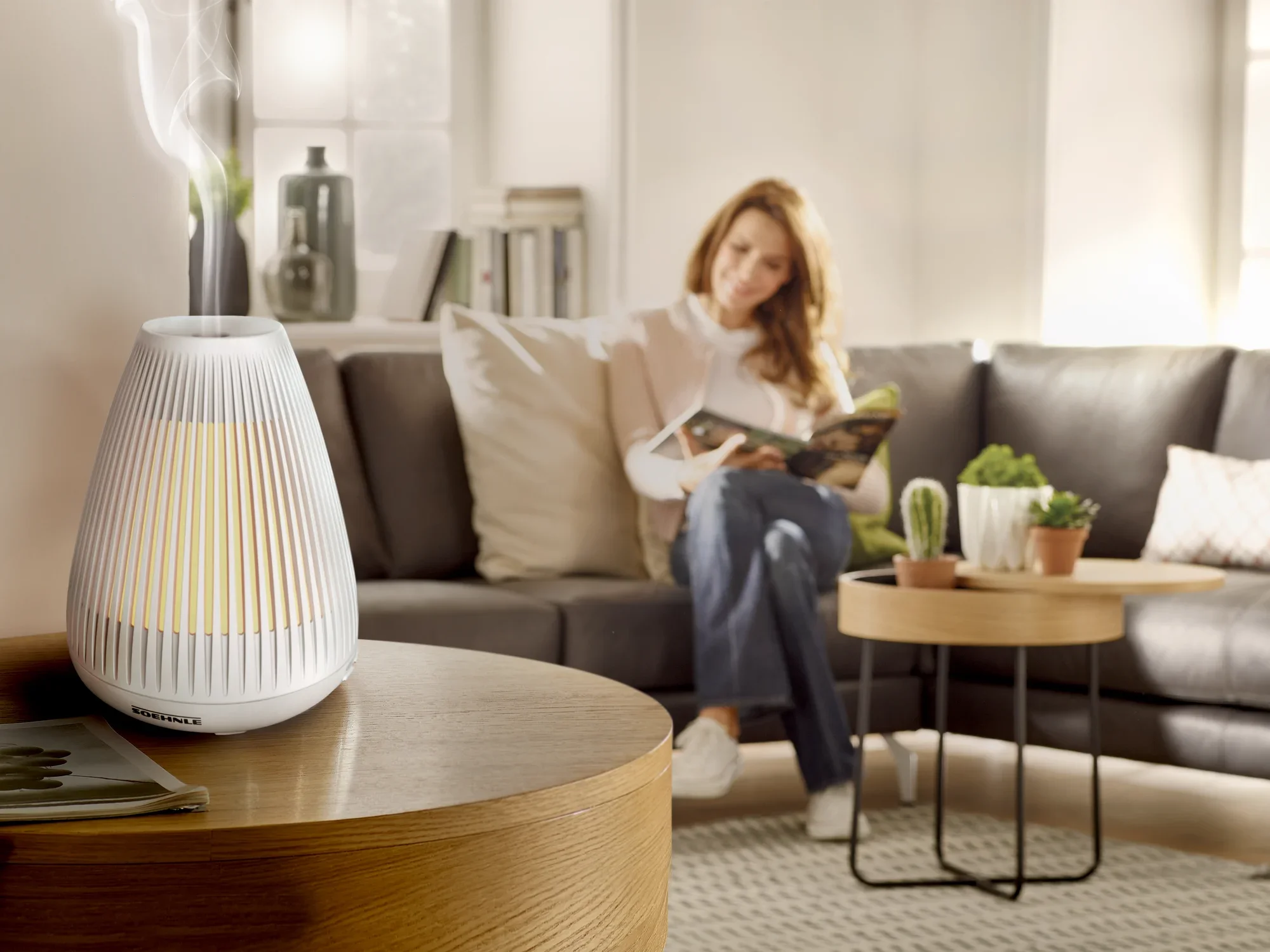 Un diffuseur d'arômes design Bari est posé sur une table en bois dans un salon confortable. À l'arrière-plan, une femme est assise sur un canapé et lit un livre.