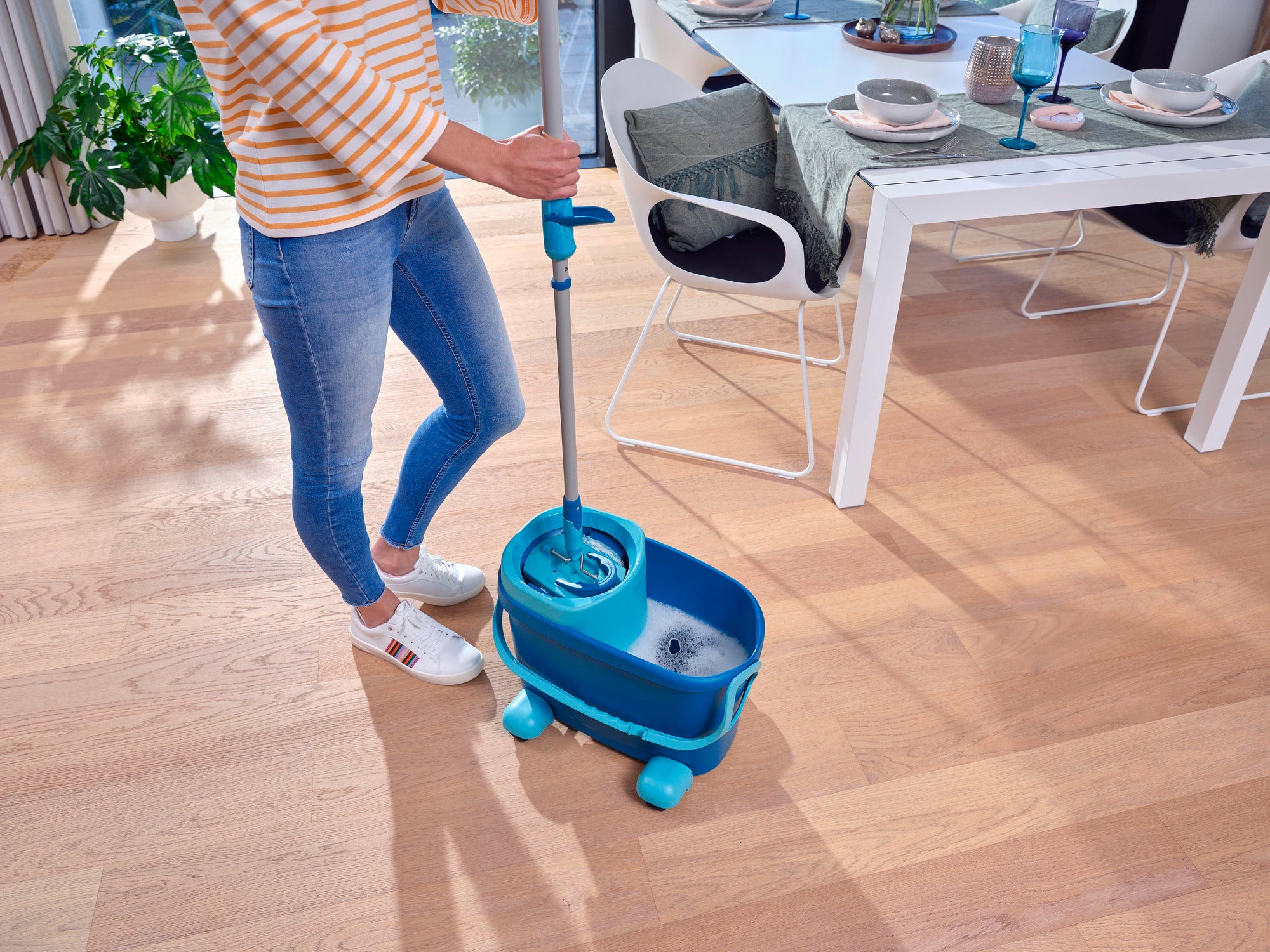 Eine Person reinigt einen Holzboden mit dem CLEAN TWIST M Ergo Set. Der Eimer hat Rollen für einfache Mobilität. Im Hintergrund ist ein gedeckter Esstisch zu sehen. Ideal für effizientes Wischen.