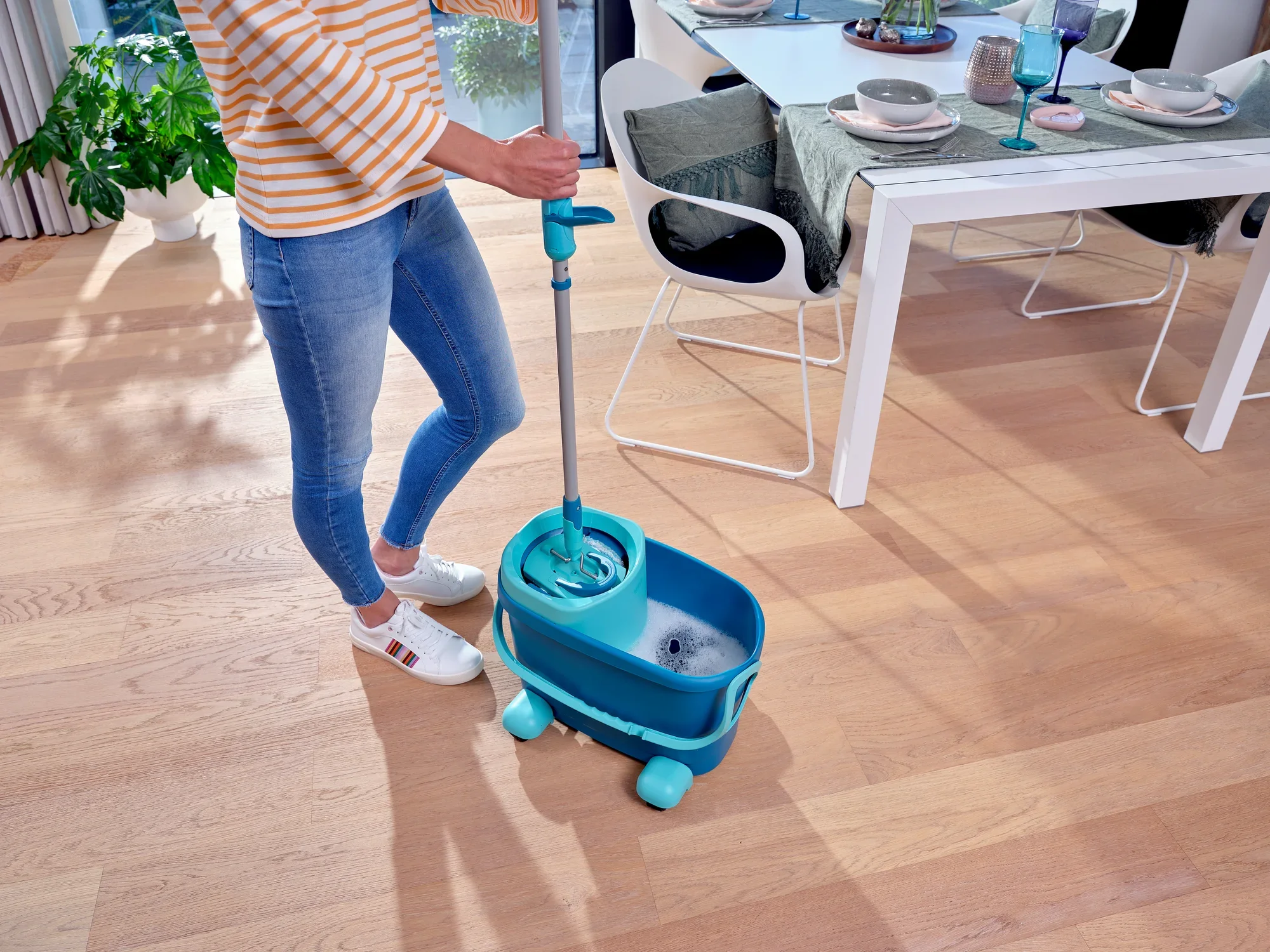 Eén persoon maakt een houten vloer schoon met de CLEAN TWIST M Ergo Set. De emmer heeft wieltjes voor eenvoudige mobiliteit. Op de achtergrond staat een gedekte eettafel. Ideaal voor efficiënt dweilen.