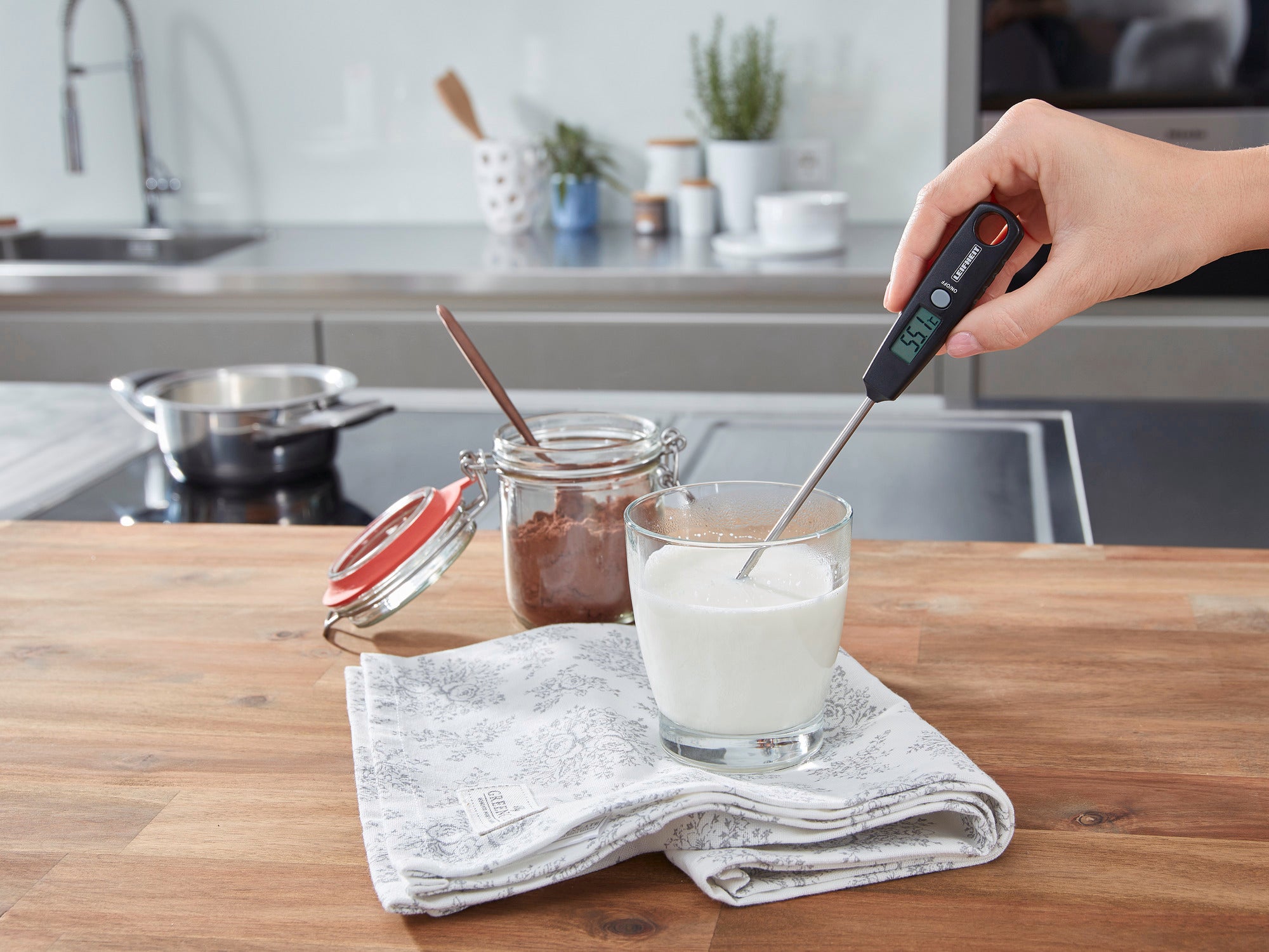 Une main mesure la temperature du lait dans un verre a l'aide d'un thermometre numerique sur une table en bois, pres d'un pot de cacao.