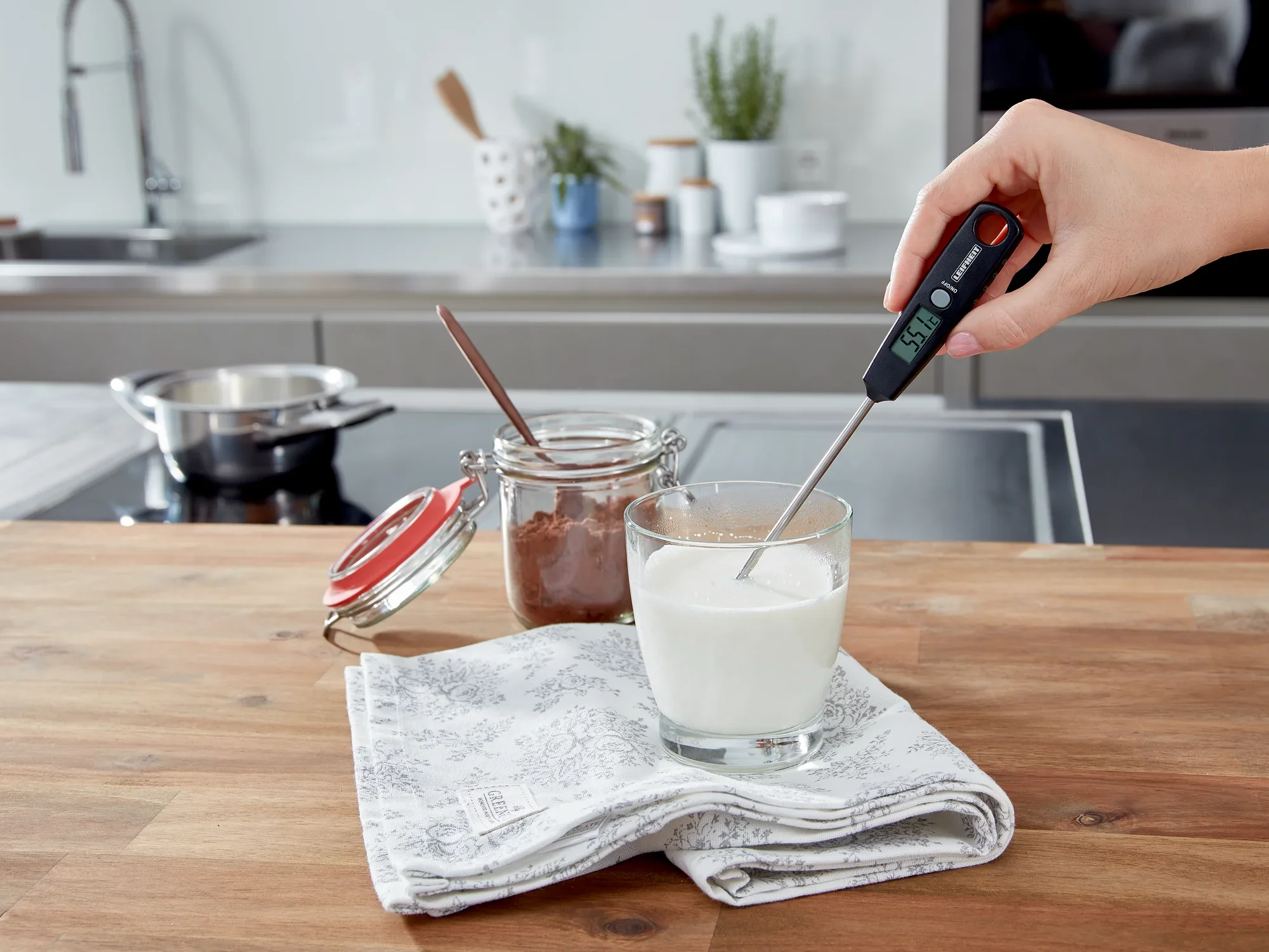 Uniwersalny termometr cyfrowy mierzy temperaturę mleka w szklance na stole kuchennym. W tle widać szklankę z łyżką i nowoczesną kuchnię.