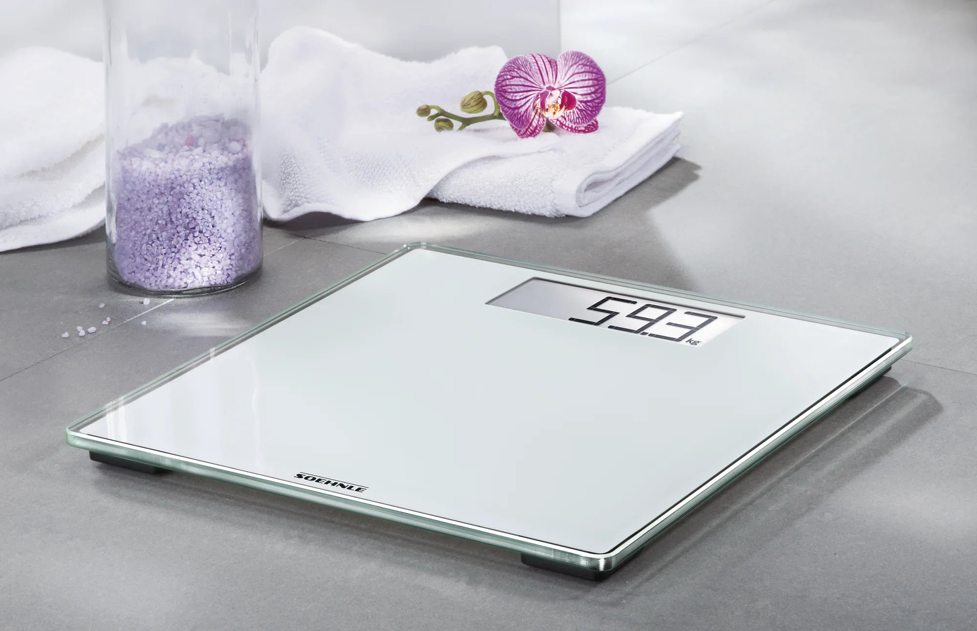 Cyfrowa waga Style Sense Comfort 100 na szarej podstawie. Waga wyświetla 59,3 kg. W tle widać ręcznik, orchideę i słoik soli do kąpieli.