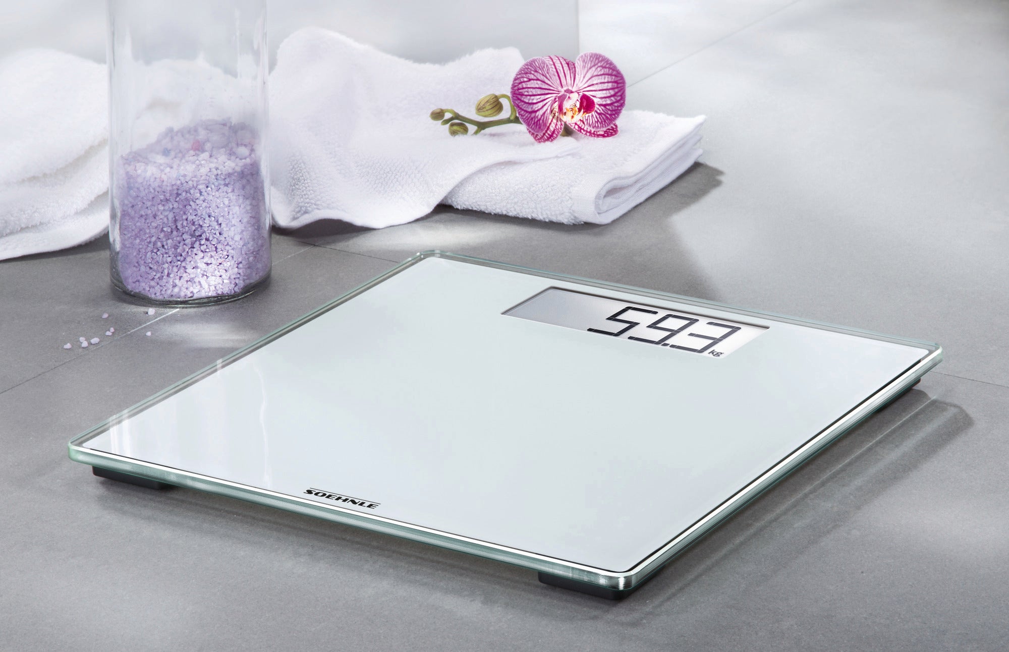 Personenweegschaal digitale Style Sense Comfort 100 op een grijze basis. De weegschaal geeft 59,3 kg aan. Op de achtergrond zie je een handdoek, een orchidee en een pot badzout.