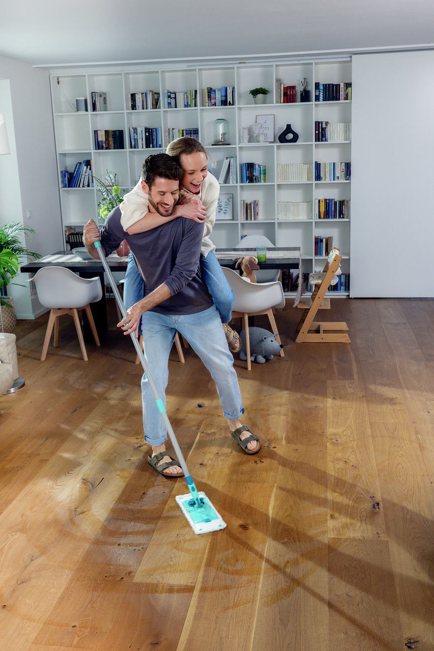 Ein lachendes Paar putzt gemeinsam den Holzboden; er wischt, waehrend sie ihn von hinten umarmt, in einem hellen Wohnzimmer.
