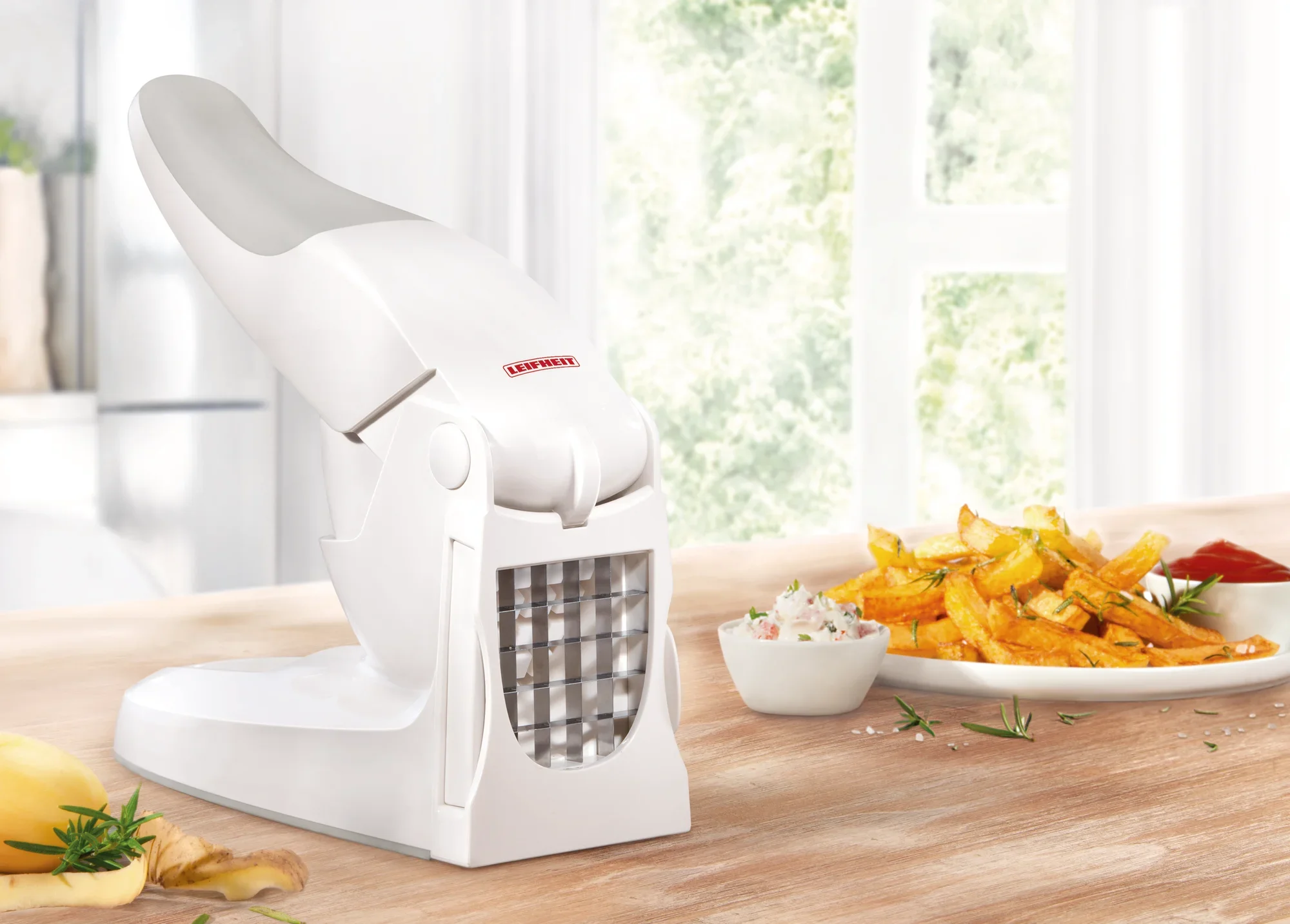 Een witte Leifheit frietsnijder staat op een houten tafel. Ernaast liggen vers gesneden frieten en een schaal met dip. Op de achtergrond zijn lichtgekleurde ramen te zien.