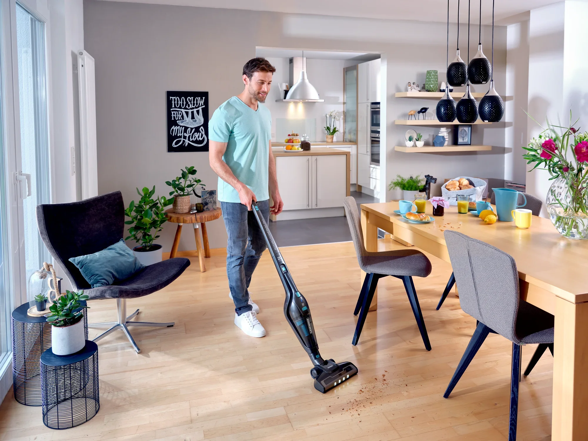 Un homme utilise l'aspirateur sans fil Rotaro PowerVac pour aspirer des miettes sur le parquet d'un salon moderne et lumineux avec coin repas. Des plantes et des décorations sont visibles à l'arrière-plan.
