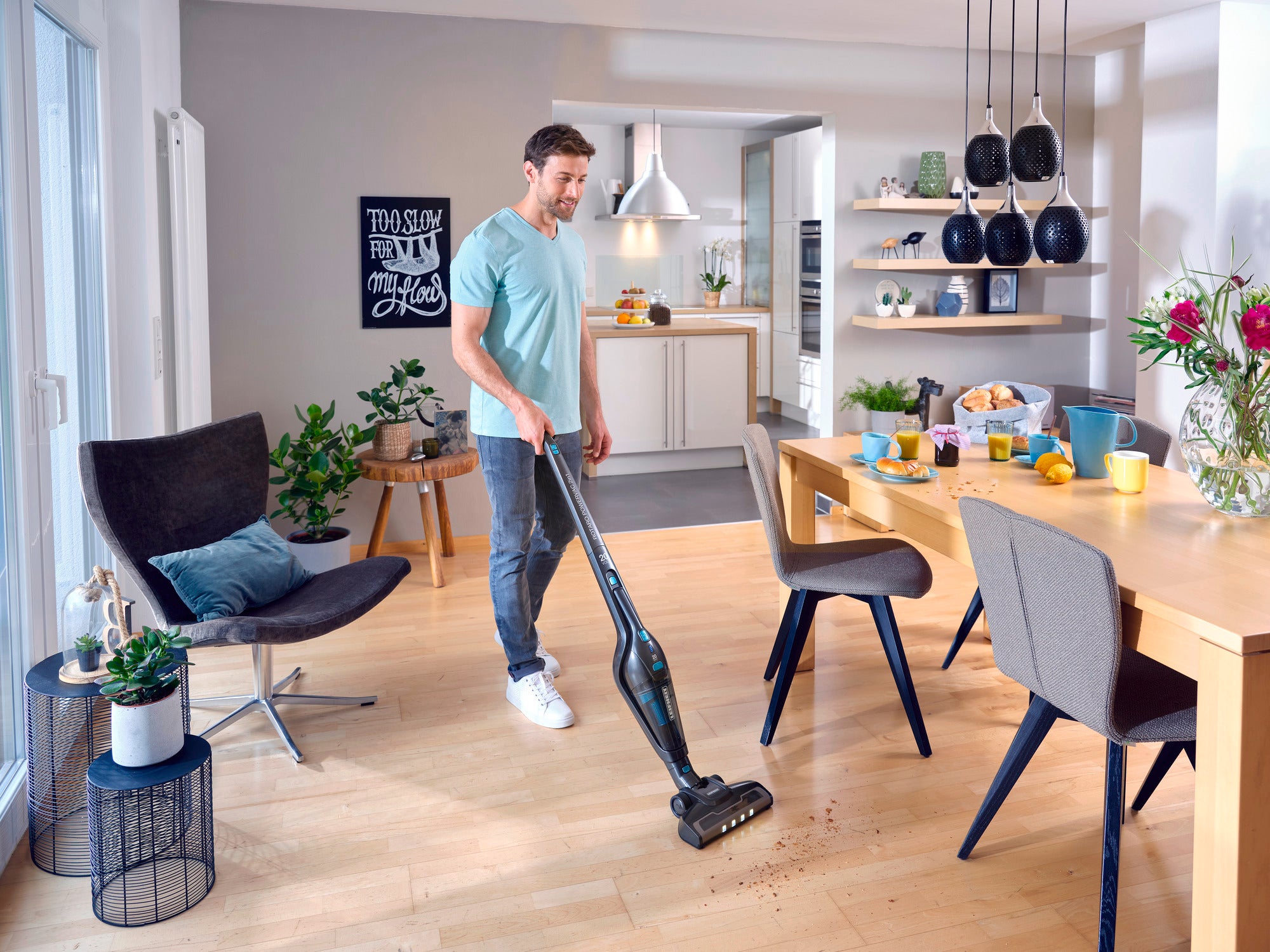 Mężczyzna używa bezprzewodowego odkurzacza Rotaro PowerVac do usuwania okruchów z drewnianej podłogi w nowoczesnym, jasnym salonie z jadalnią. W tle widoczne są rośliny i dekoracje.