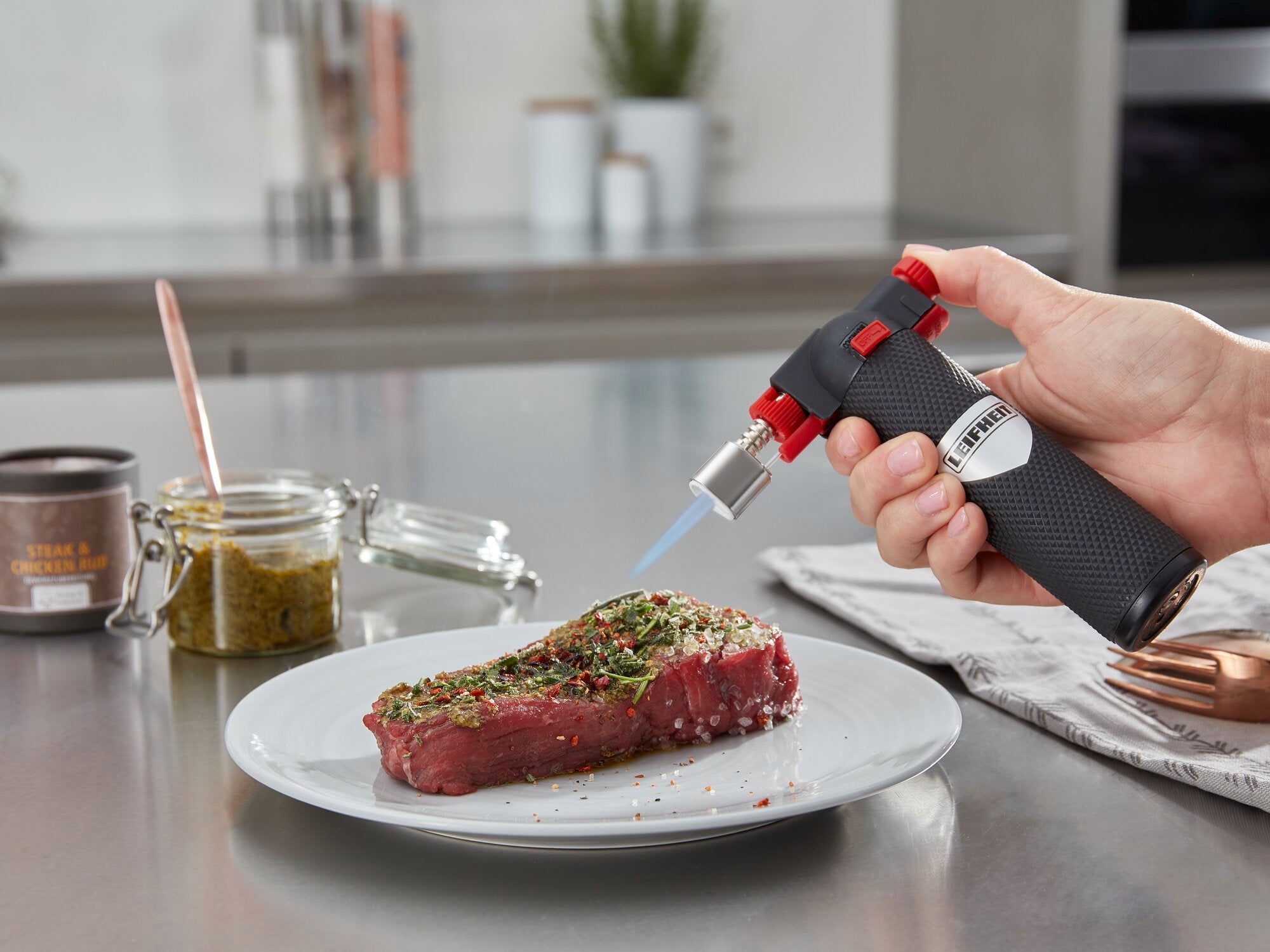 Een flammkuip wordt gebruikt om een gekruide steak op een bord te flamberen. Kruiden en keukengerei zijn te zien op een werkblad op de achtergrond.