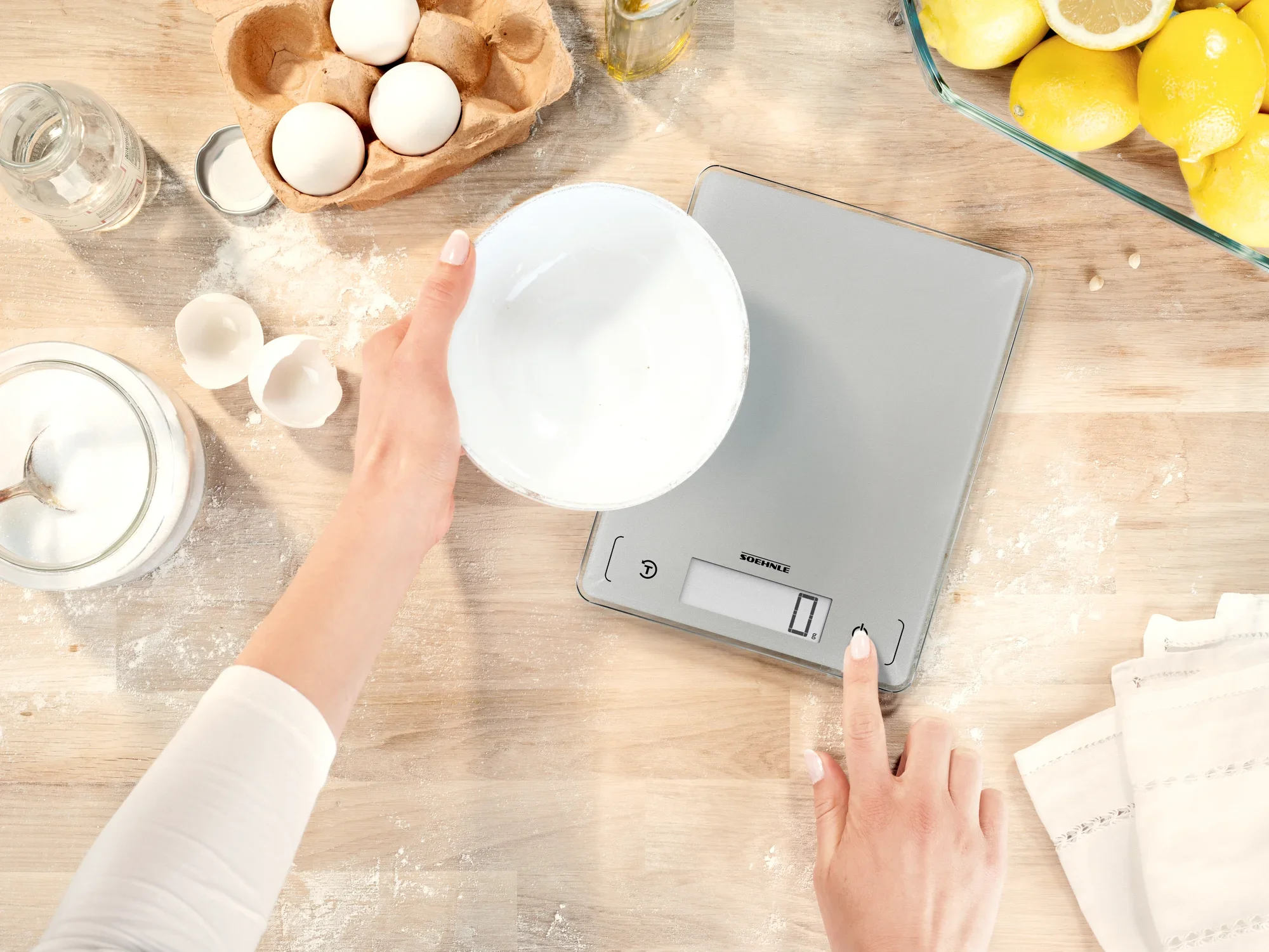 Een persoon weegt ingrediënten in een kom op een digitale keukenweegschaal, model Page Comfort 300 Slim. De weegschaal rust op een houten oppervlak, omringd door eieren en citroenen.