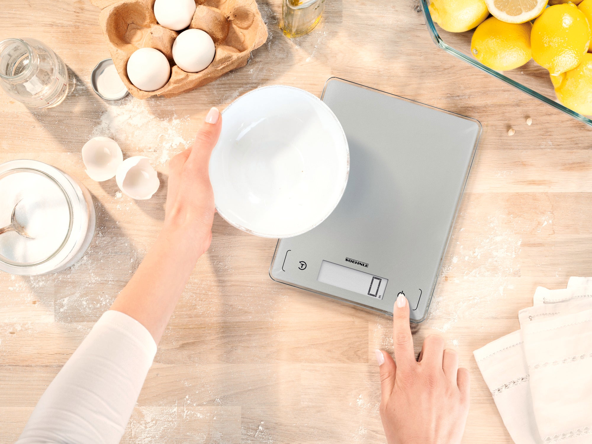 A person weighs ingredients in a bowl on a Page Comfort 300 Slim digital kitchen scale. The scale rests on a wooden surface surrounded by eggs and lemons.