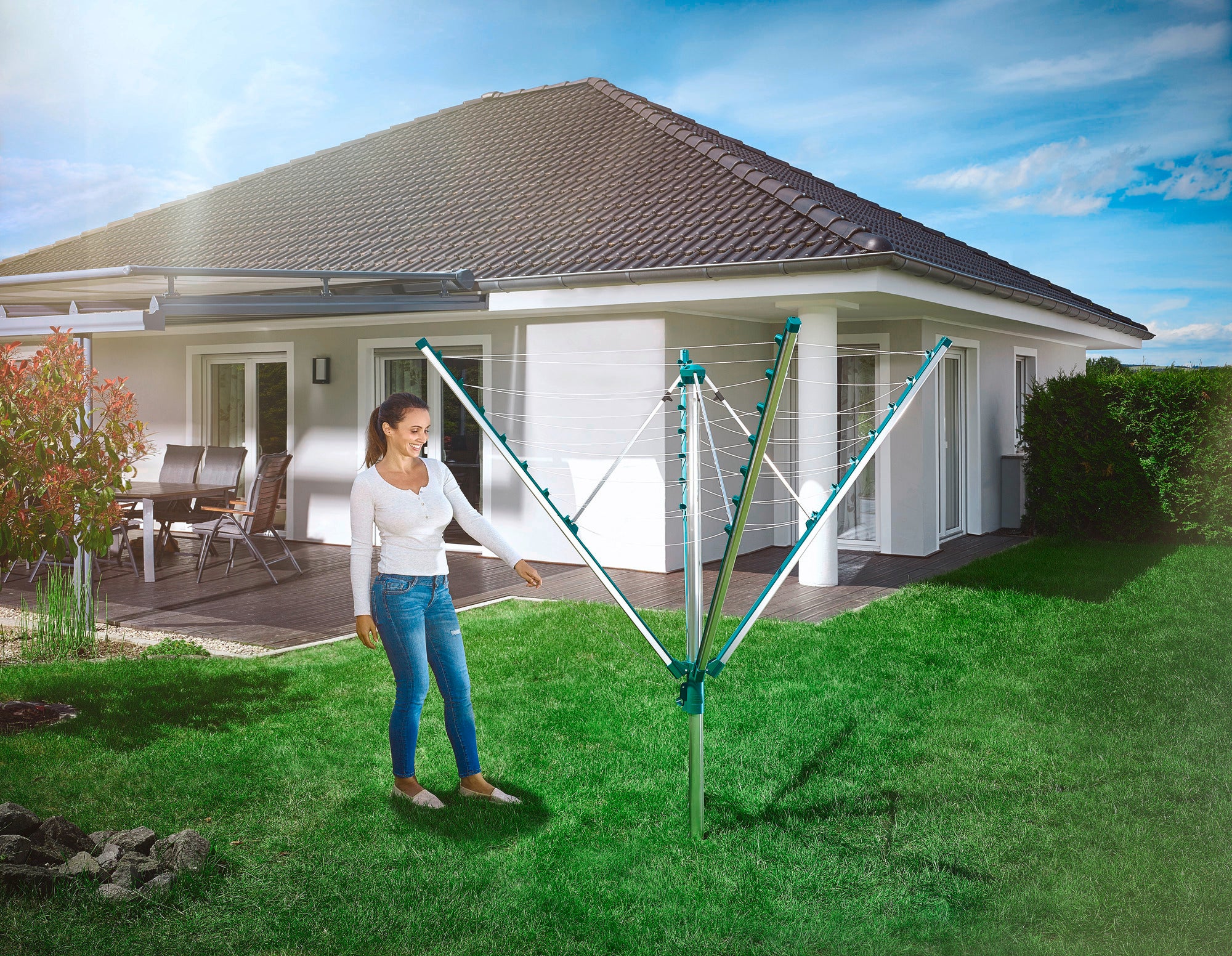 Eine Frau steht im Garten neben einer geöffneten Leifheit Linomatic 400 Comfort Wäschespinne. Im Hintergrund ist ein modernes Einfamilienhaus mit Terrasse zu sehen.