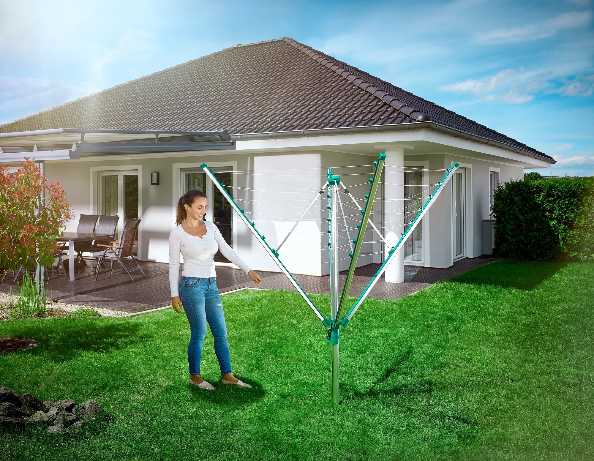 Une femme se tient dans le jardin à côté d'un séchoir à linge Leifheit Linomatic 400 Comfort ouvert. À l'arrière-plan, on aperçoit une maison individuelle moderne avec une terrasse.