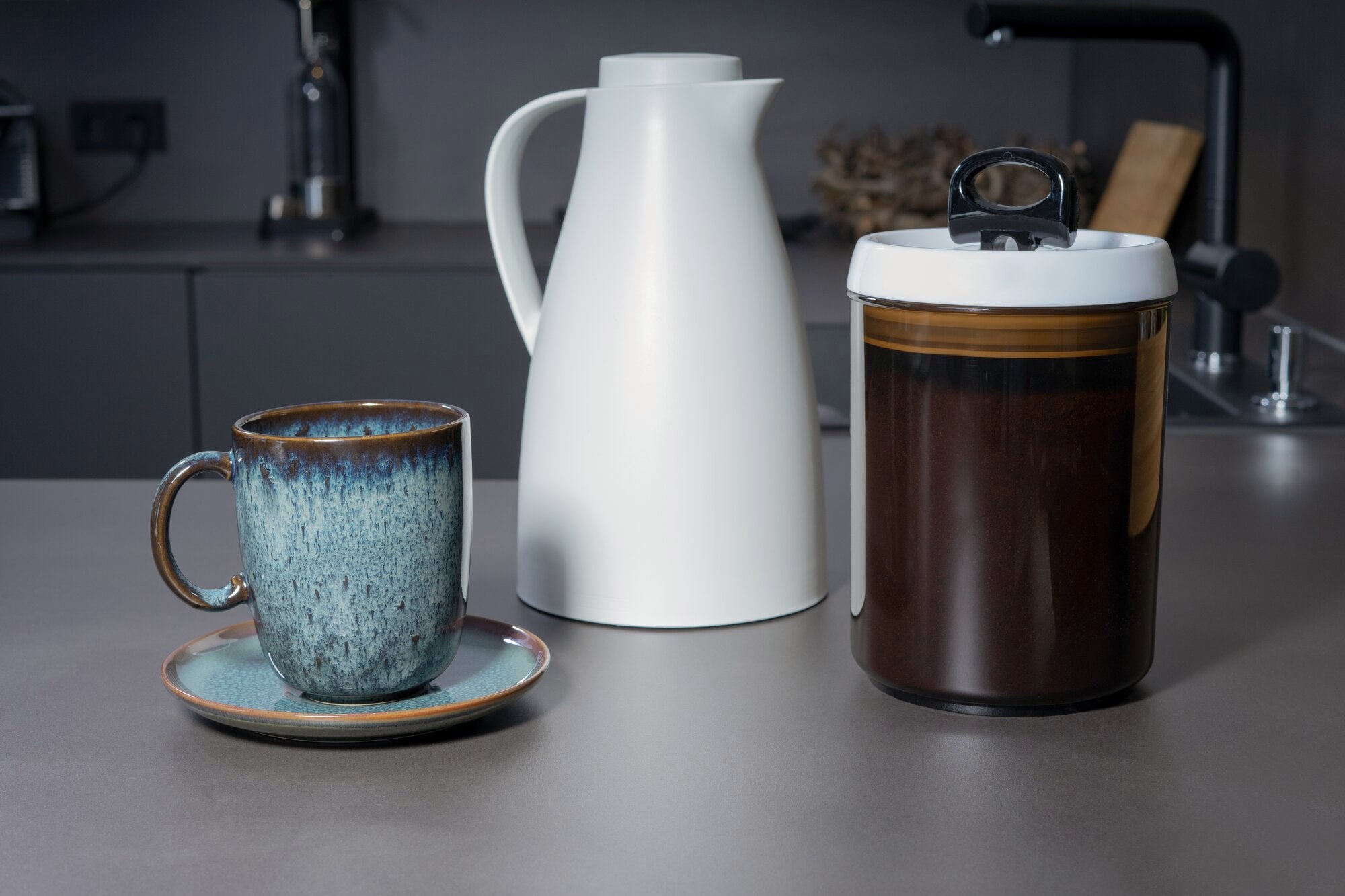 Ein Vorratsbehälter "Fresh and Easy" für Kaffee steht auf einer Küchenarbeitsplatte. Daneben befinden sich eine weiße Kanne und eine blaue Tasse auf einem Unterteller.