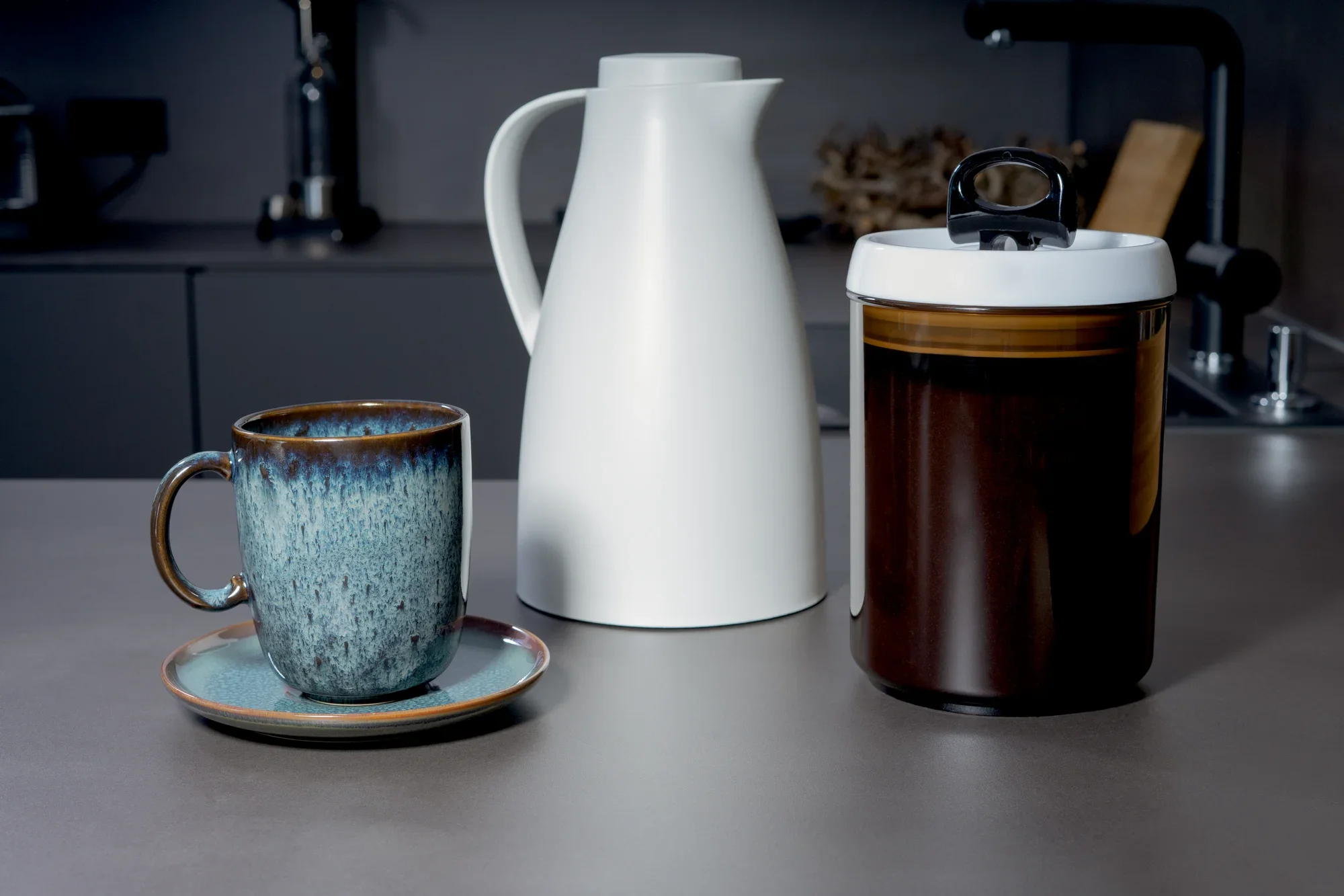 Pojemnik do przechowywania kawy "Fresh and Easy" stoi na blacie kuchennym. Obok stoi biały dzbanek i niebieska filiżanka na spodku.